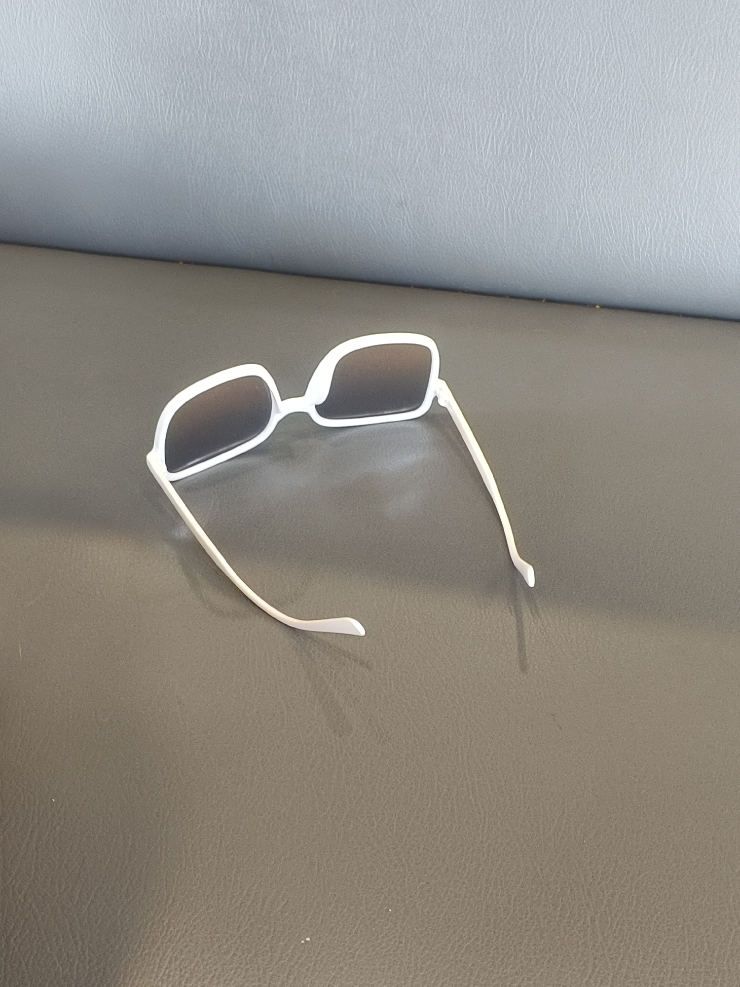A pair of white-rimmed sunglasses with dark lenses is positioned on a textured gray surface, possibly a bench or a couch. The sunglasses are slightly angled, with one arm extended outwards. The background consists of a light gray, textured surface that appears to be a wall or the back of the furniture. The scene is lit from above and to the left, casting a slight shadow from the sunglasses onto the gray surface. There are no people visible in the image. The location is not discernible from the image content alone and cannot be placed in Denpasar, Indonesia.