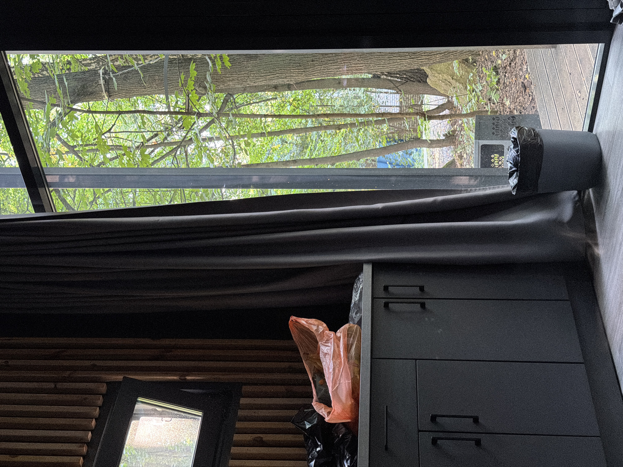 The image depicts an interior scene looking out a large window at a wooded area. In the foreground, a dark gray cabinet with black handles is visible, with a countertop above it. On the countertop, there are several plastic bags, one of which is orange and appears to be filled with items. To the right of the cabinet, a gray trash can with a black liner is positioned. The large window reveals a dense green forest with several trees. The ground outside is covered with leaves and branches, and a wooden deck is partially visible. The lighting suggests it is daytime, possibly overcast or late afternoon, as the light is diffused. The overall impression is one of a somewhat cluttered domestic or utility space adjacent to a natural environment. The location is not specified in the image.