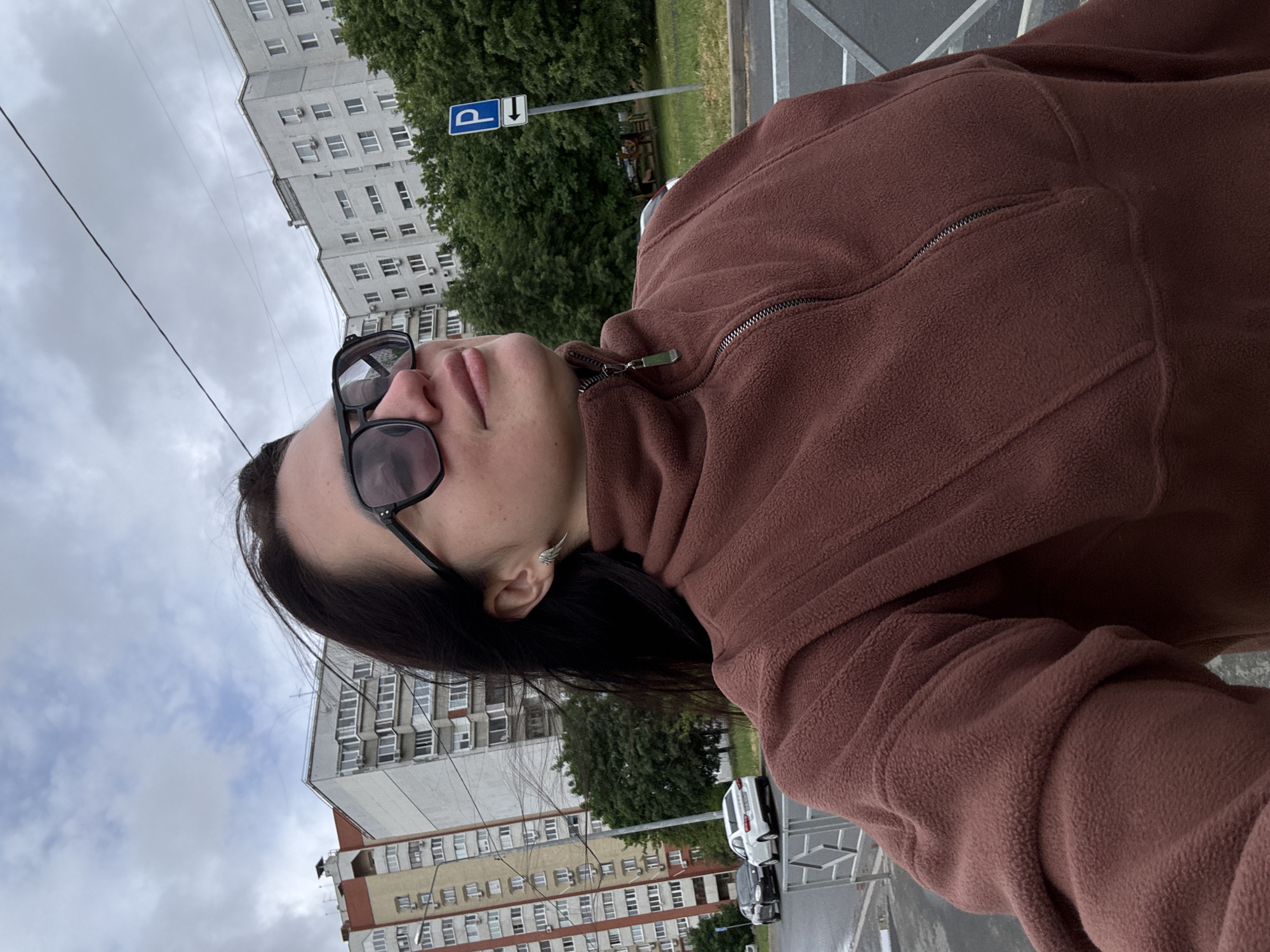 A selfie shows a person with dark hair wearing sunglasses and a brown fleece jacket. In the background are buildings and trees. A parking sign is also visible. The picture appears to be taken in an urban setting.