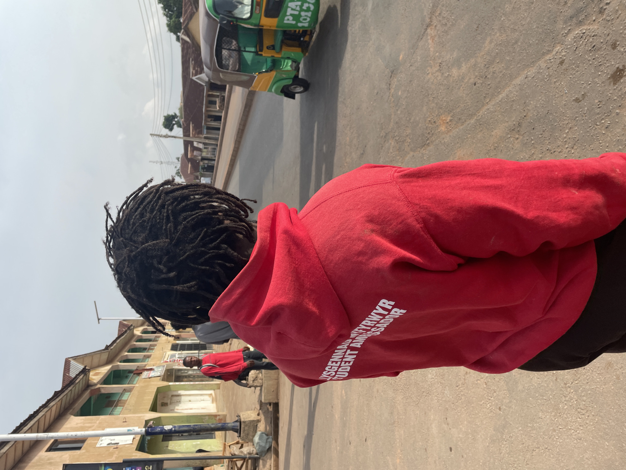 The image shows a person with dreadlocks wearing a red hoodie with white text that reads "LYSGENNAD WEYFRWYR STUDENT AMBASSADOR". They are standing on a street in what appears to be Barakin Lerawi, Nigeria. A green and yellow "keke" (auto rickshaw) with "PTA 10136" visible on the front is in the background on the right side of the image. Several buildings are visible on the left side of the image, as well as a man in a red shirt walking away from the camera in the distance. The environment appears to be an urban street with a mix of residential and commercial buildings. The ground is paved with asphalt. The weather seems to be clear and bright.