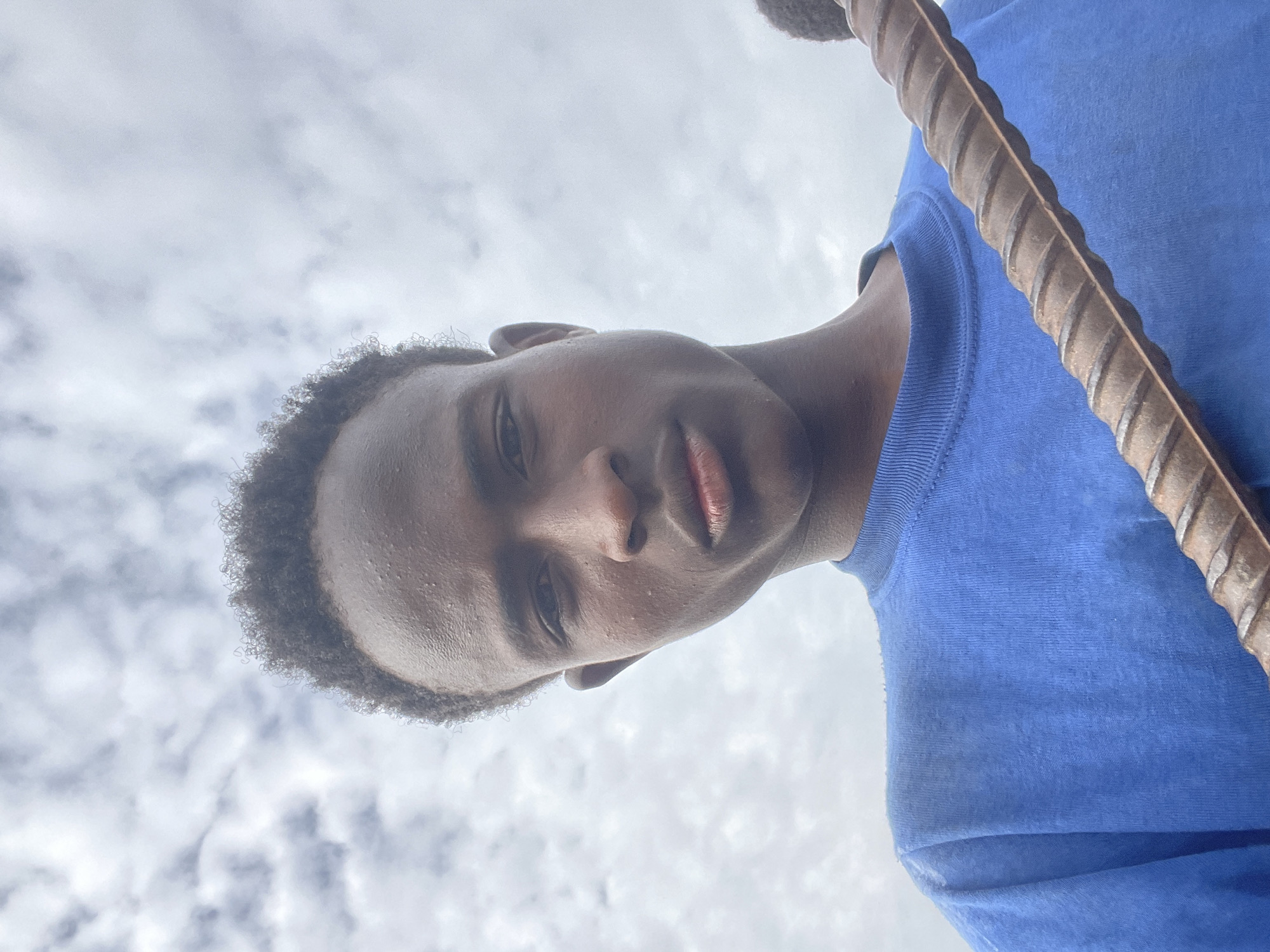A young Black male is shown from a low angle, looking up at the camera. He has dark skin, short, curly dark hair, and is wearing a bright blue t-shirt. A rusty, ridged metal bar is held diagonally across the lower right portion of the frame, obscuring part of the man's body. The background is a cloudy sky. The image is likely taken outdoors in Jos, Nigeria.