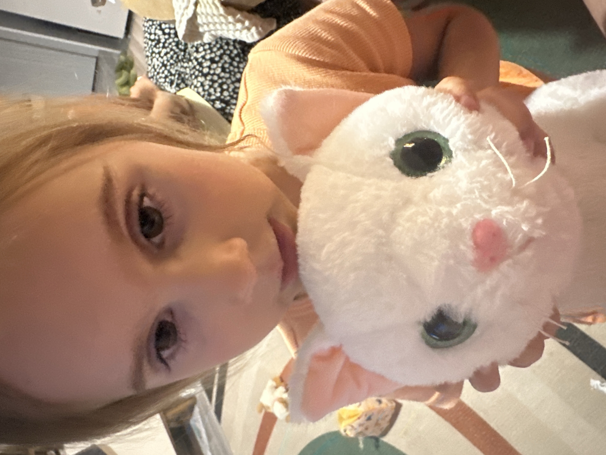 A fair-skinned child with light brown hair is holding a white plush cat with green eyes and a pink nose. The child wears an orange top. The background includes a black and white patterned garment, a table, and a floor with a rug. The location is indeterminable.