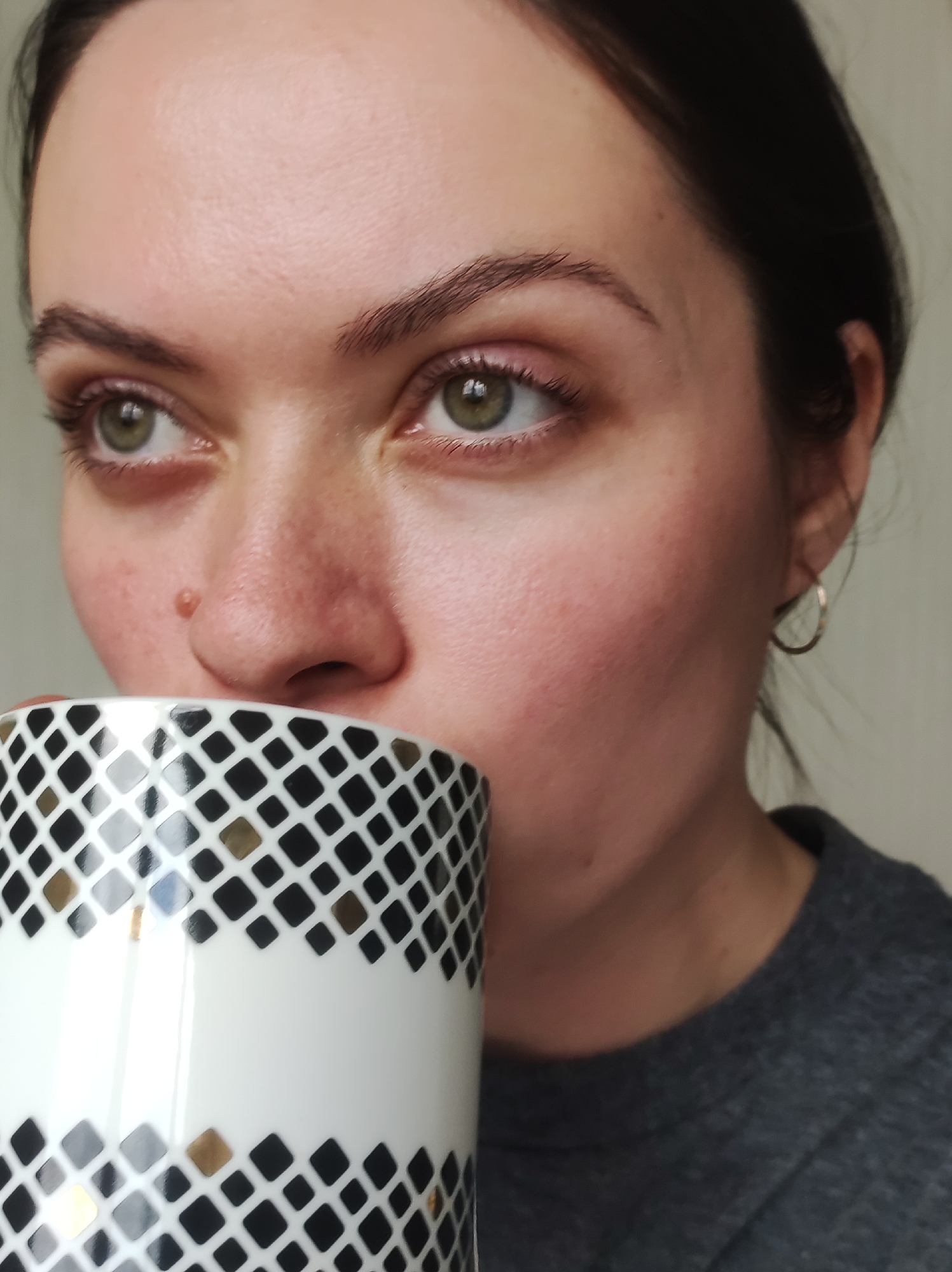 A fair-skinned woman with dark hair pulled back is shown from the chest up, holding a white mug with a black and gold geometric pattern to her lips. Her eyes are green, and she has a mole on the side of her nose. She wears small hoop earrings and a gray t-shirt. The background is a plain, light-colored wall. There is no information in the image to suggest the location is in Chaikovsky, Russia.