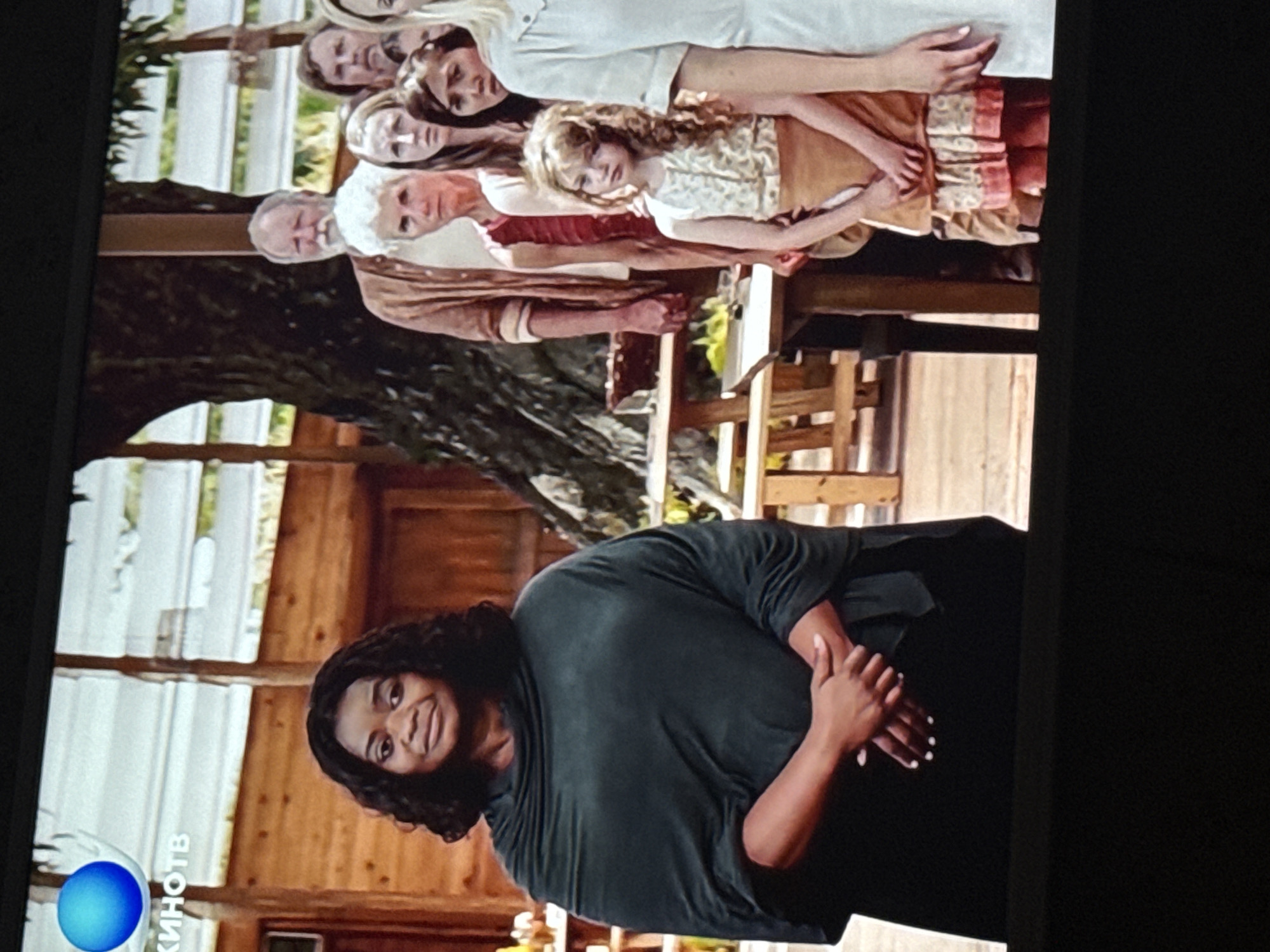 A horizontal image depicts two scenes. The bottom scene features a Black woman wearing a dark, draping garment. She has short, curly hair and is smiling, with her hands clasped in front of her. The background appears to be the interior of a wooden cabin. The upper scene shows a group of people in various ages, dressed in vintage clothes. They seem to be standing behind a table. The backdrop consists of greenery, possibly a garden or outdoor scene. The image is displayed on a screen that has the logo of "KинoТВ" in the bottom left corner. The location is Mytishchi, Russia.