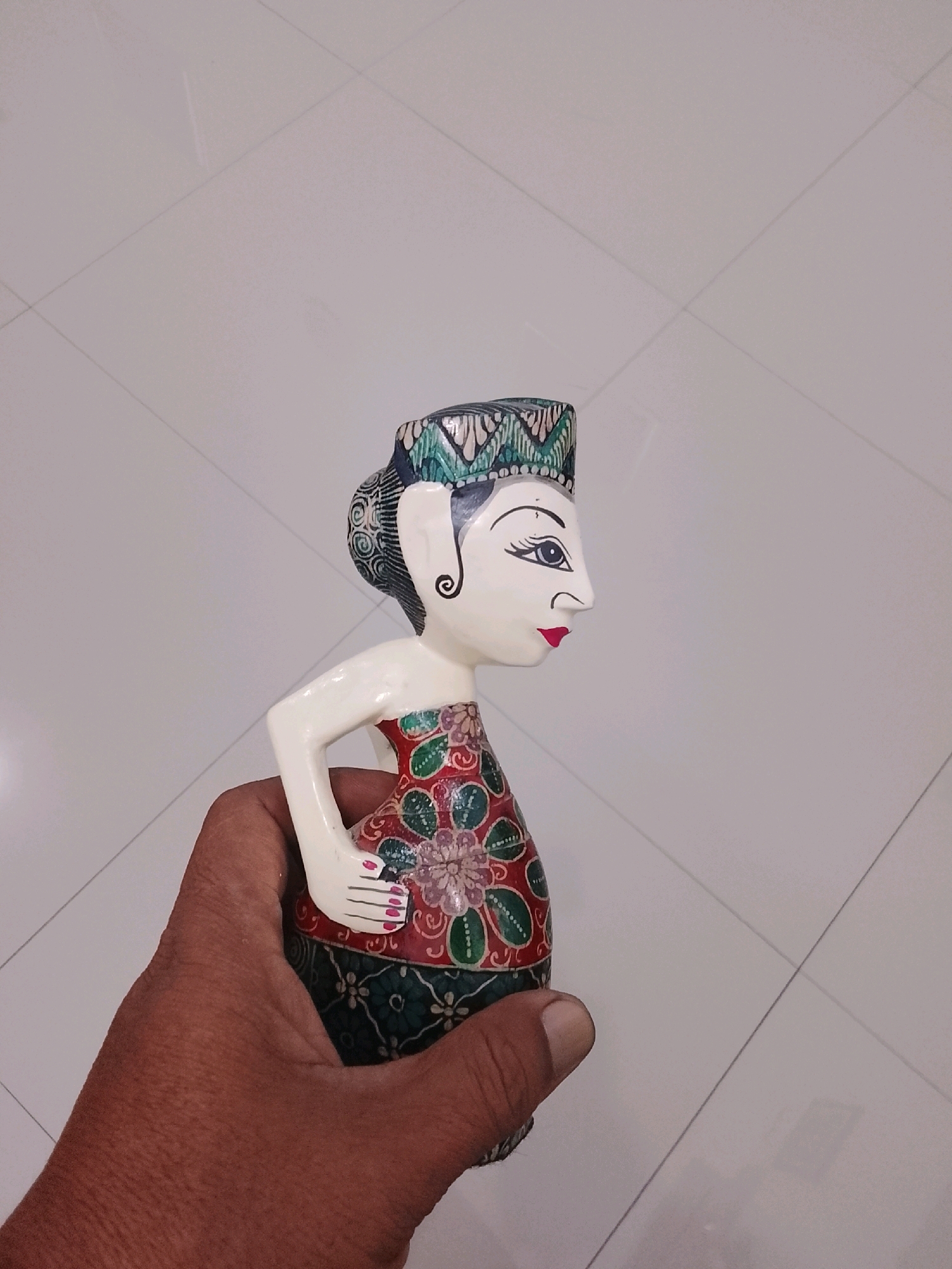 A hand is holding a traditional Javanese wooden puppet, known as a wayang golek, in Slawi, Indonesia. The puppet is brightly painted with a patterned headdress, a white face with dark eyes and red lips, and a red and green patterned garment. The background is a light-colored tiled floor.