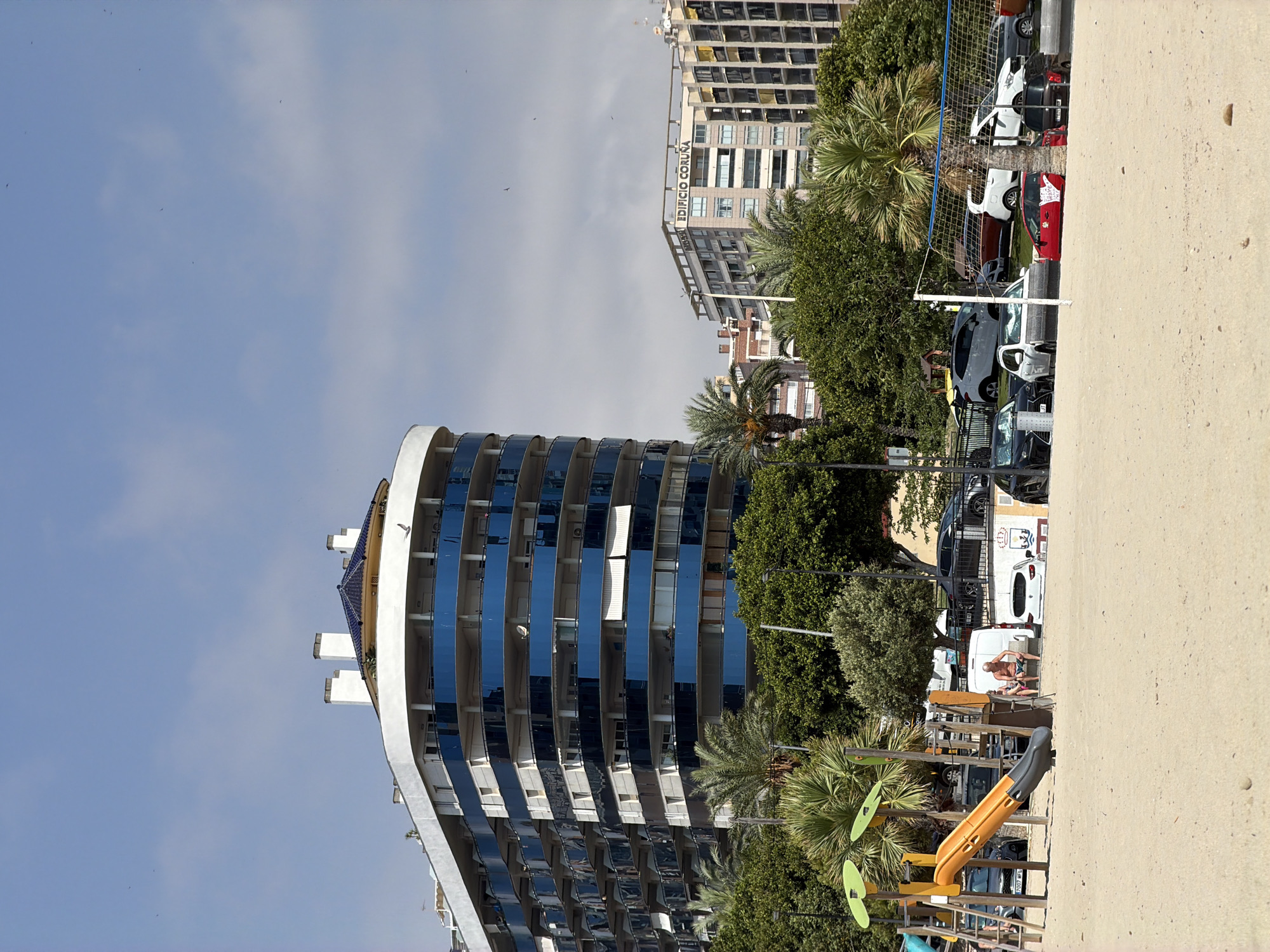 This media file shows an outdoor scene with a beach in the foreground, a playground, trees, buildings, and parked cars in the background. The image is oriented vertically, with the sky at the top and the beach at the bottom.
In the foreground, there is a sandy beach with a children's playground featuring a yellow slide and green leaf-shaped decorations. Beyond the playground, there are several trees, including palm trees.
In the middle ground, there are several parked cars, including white vans and other vehicles. A white van has a drawing on its side. Two people are sitting on a low wall near the vans. A volleyball net is visible.
In the background, there are modern buildings. The most prominent building is a curved structure with many floors and reflective blue windows. Further in the background, there is a taller, older building with many windows, some of which appear empty or damaged. The text "EDIFICIO CORUÑA" is visible on this building.
The sky is blue with some clouds. The lighting suggests it is daytime, likely sunny. The overall setting appears to be a coastal urban area, possibly a resort, in Spain, as indicated by the location context. The scene conveys a sense of leisure and urban life.