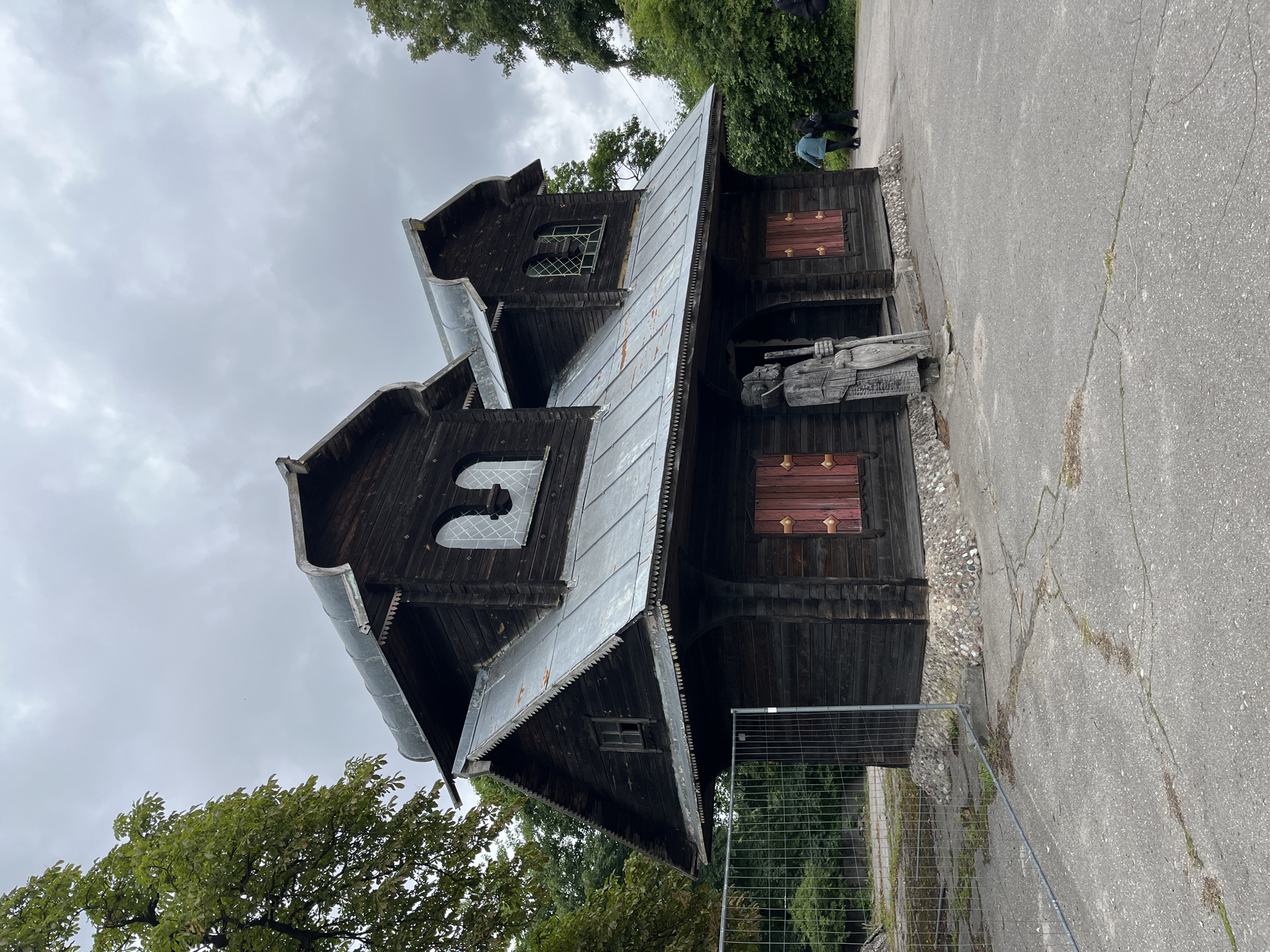 The image depicts an old, dark wooden house with a metal roof, reminiscent of traditional Russian architecture. The house has two stories, with arched windows on the upper level and smaller, square windows on the lower level. Red wooden shutters are visible on some of the lower windows. A large wooden sculpture of a figure holding a staff stands outside the entrance. The house is surrounded by trees and a gray, cracked asphalt surface. A metal fence is visible in the foreground, and two people are walking in the background on the right side of the image. The sky is overcast. This structure is identified as the "House of Fishermen" in the Museum of wooden architecture in Kaliningrad, Russia.