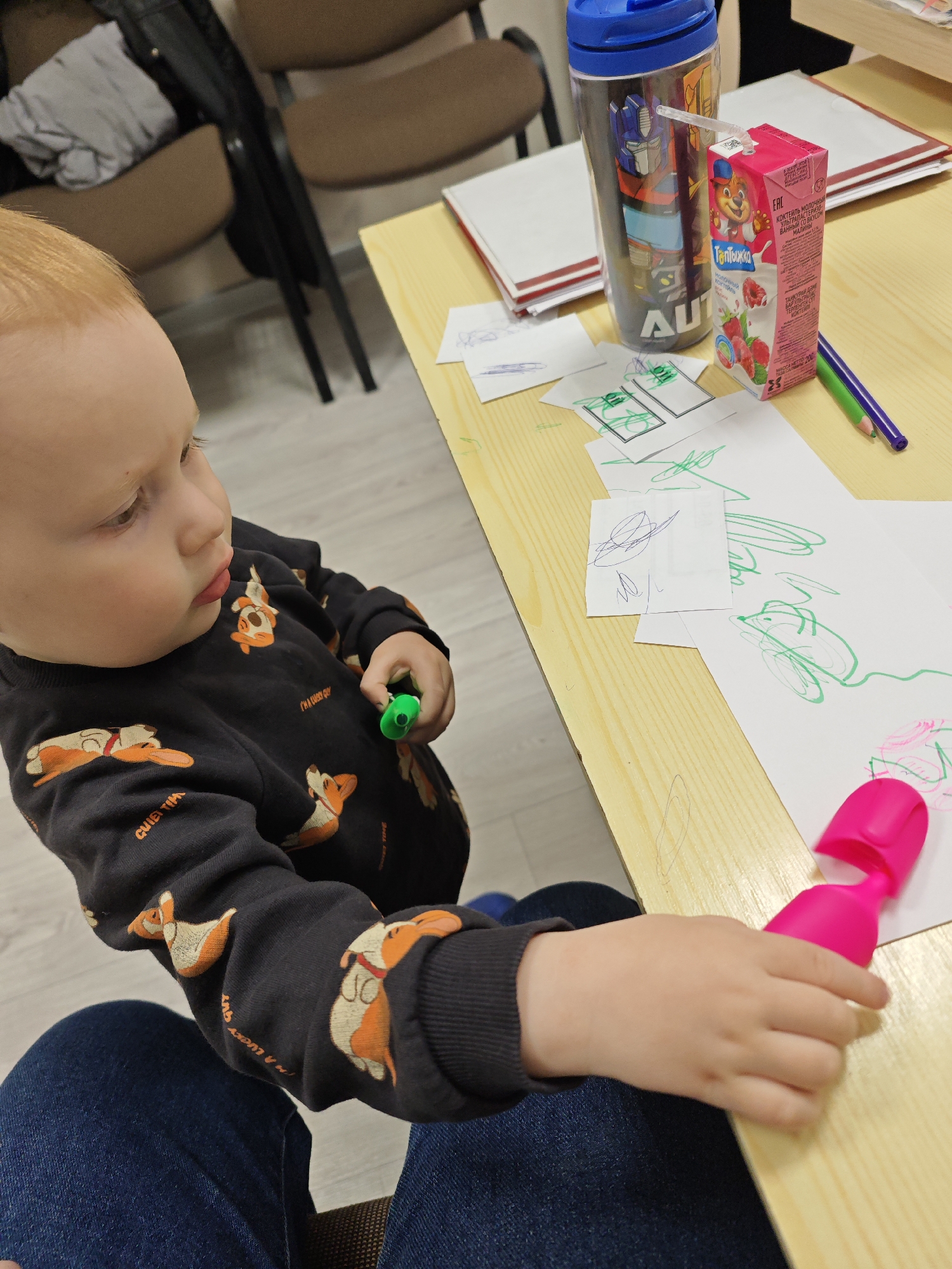 In an indoor setting in Markovskii, Russia, a young child with reddish-blonde hair is engaged in drawing. The child is seated on an adult's lap, seen wearing dark jeans, and is focused on the paper in front of them. Their left hand holds a small green marker, while their right hand is near a larger, bright pink marker.
The child is wearing a black long-sleeved top patterned with orange and white cartoon corgi-like dogs. Visible text on the shirt includes "I'M A LUCKY GUY" and "QUIET TIME".
The activity takes place at a light wooden table, which is cluttered with art supplies and drinks. Several sheets of white paper display green and pink scribbles, indicating active drawing. Other items on the table include a blue water bottle with an "AUTOBOTS" logo and partially visible text "AUT", a small pink juice box with a cartoon bear character, and green and purple colored pencils.
The juice box packaging features Russian text: "Топтыжка" (Toptyzhka, likely the brand name or character's name), "молочный коктейль" (milk cocktail), and "УЛЬТРАПАСТЕРИЗОВАННЫЙ СО ВКУСОМ МАЛИНЫ" (UHT with raspberry flavor), along with "EAC" certification. The child appears calm and absorbed in their creative endeavor. In the background, two brown office-style chairs are visible.