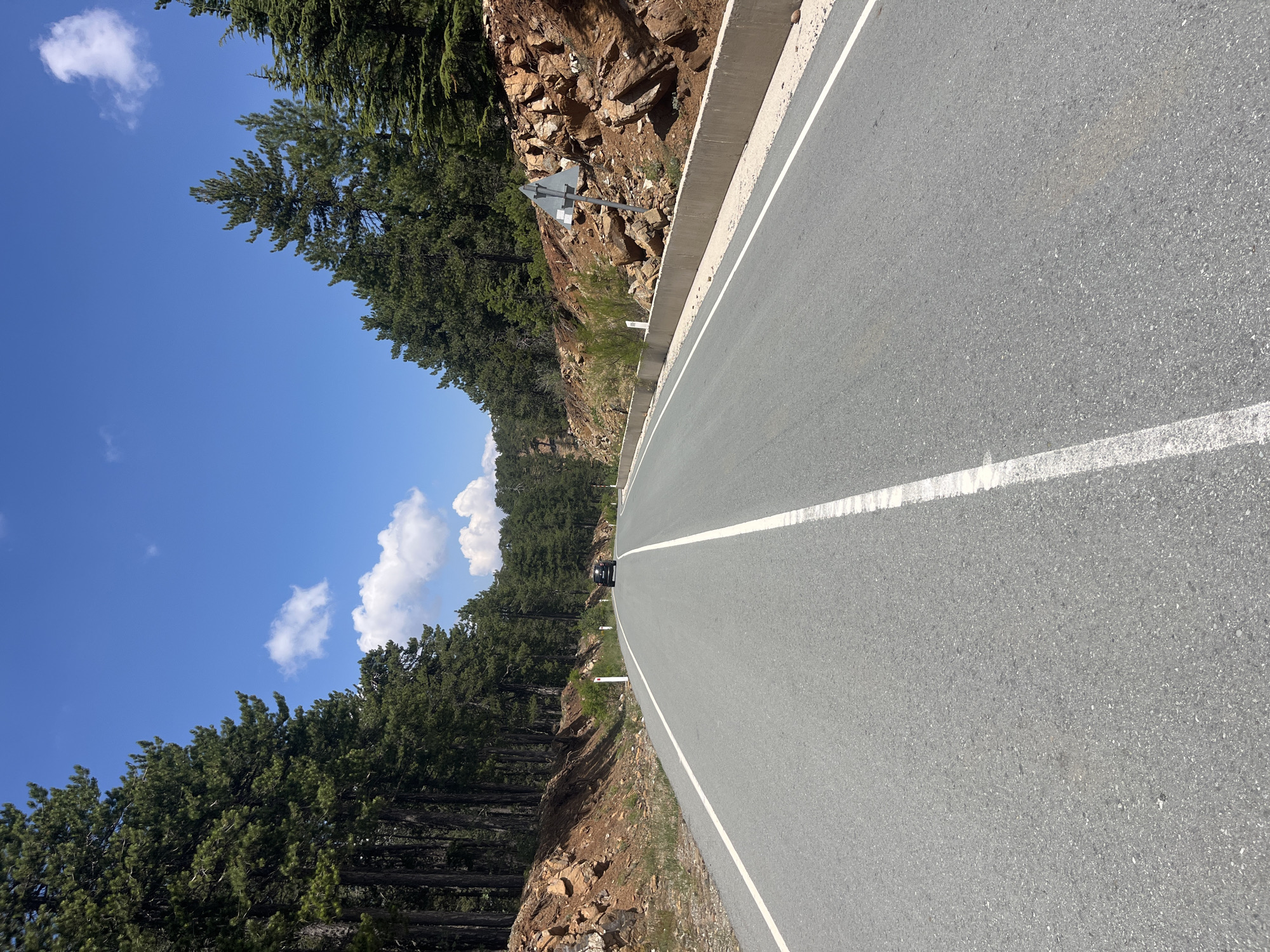 The image depicts a road through a pine forest in Troodos, Cyprus. A dark-colored car is visible driving away from the viewer on the asphalt road, which is marked with a white dashed line in the center and a solid white line on the right edge. The road is flanked by a steep, rocky embankment covered in brown soil and scattered stones on the right, and a dense forest of tall pine trees on the left. A concrete barrier runs along the edge of the embankment. The sky is clear blue with a few scattered white clouds. The scene suggests a sunny day with good visibility.