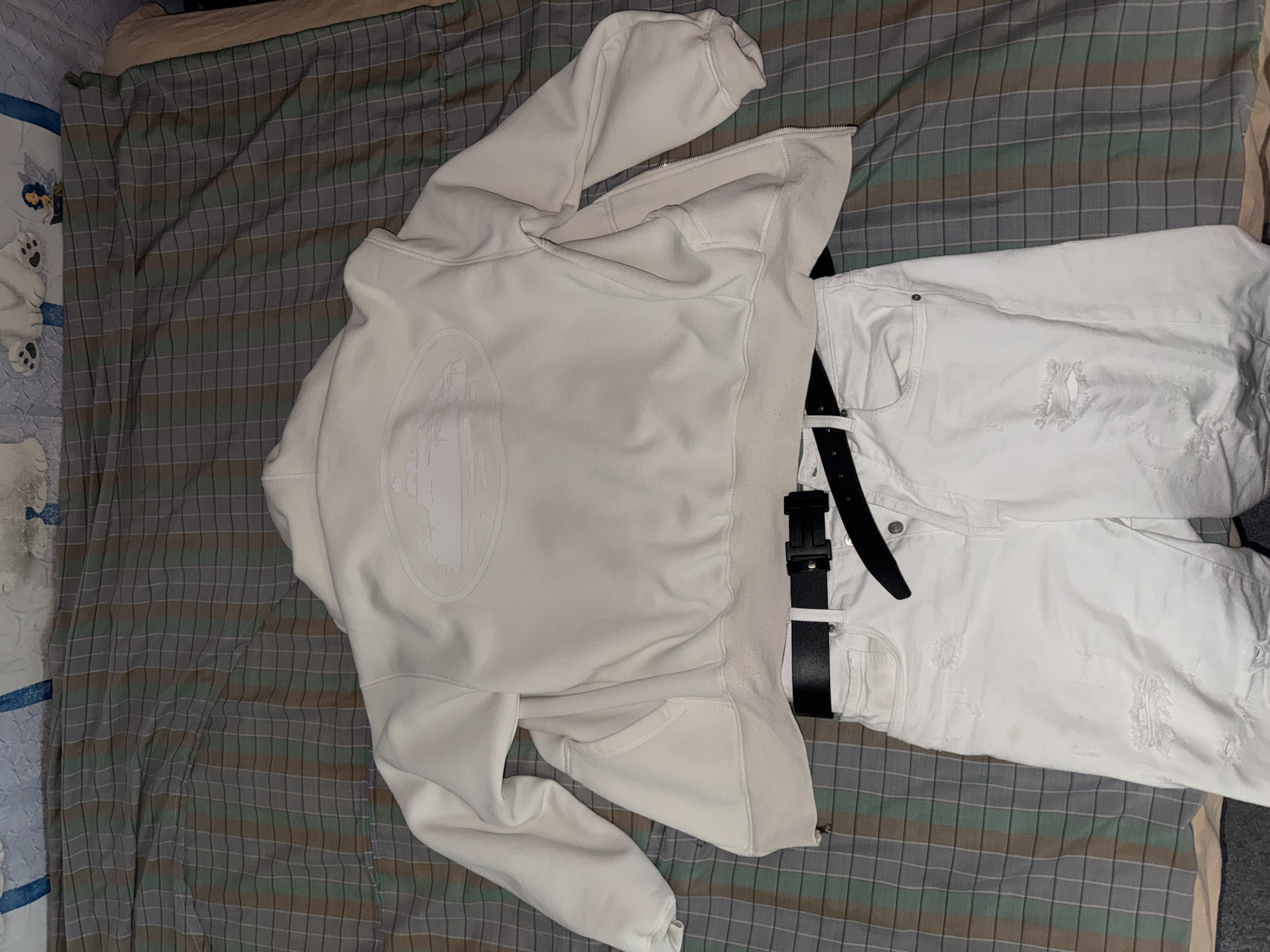 The image depicts a white hooded sweatshirt with a graphic on the back, paired with white distressed jeans and a black belt. The items are arranged on a bed with a striped bedsheet and another patterned blanket partially visible. The location is not discernible and there is no indication that the photo was taken in Ruse, Bulgaria. There are no people, actions, or interactions visible in the image.