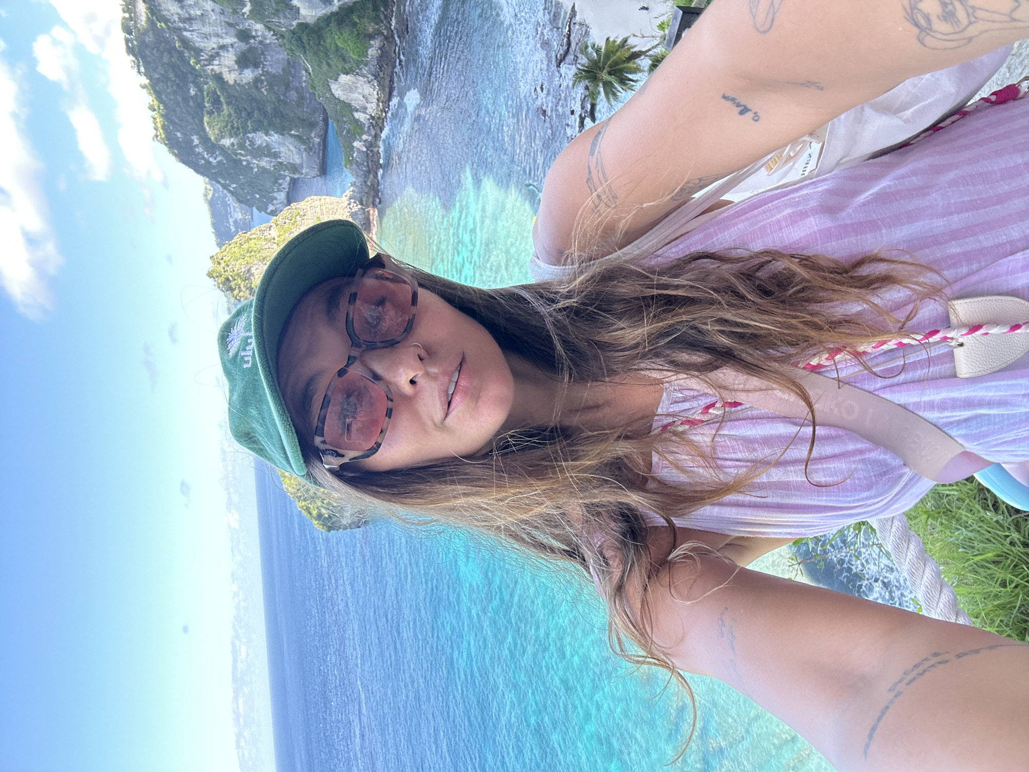 A young woman is pictured in a selfie, her face partially obscured by sunglasses and a green baseball cap. She has brown hair, fair skin, and a nose ring. She is wearing a light pink and white striped shirt with a bag slung over her shoulder. Tattoos are visible on both of her arms.
In the background, there is a stunning coastal view featuring clear turquoise water. A rocky cliff formation is visible on the left side of the frame, and further back, another green, rocky outcrop juts into the ocean. The sky is light blue with some clouds. Some green vegetation is at the bottom.
Based on the landscape, the photo appears to have been taken in Nusa Penida, Indonesia, an island known for its scenic coastlines and rock formations.