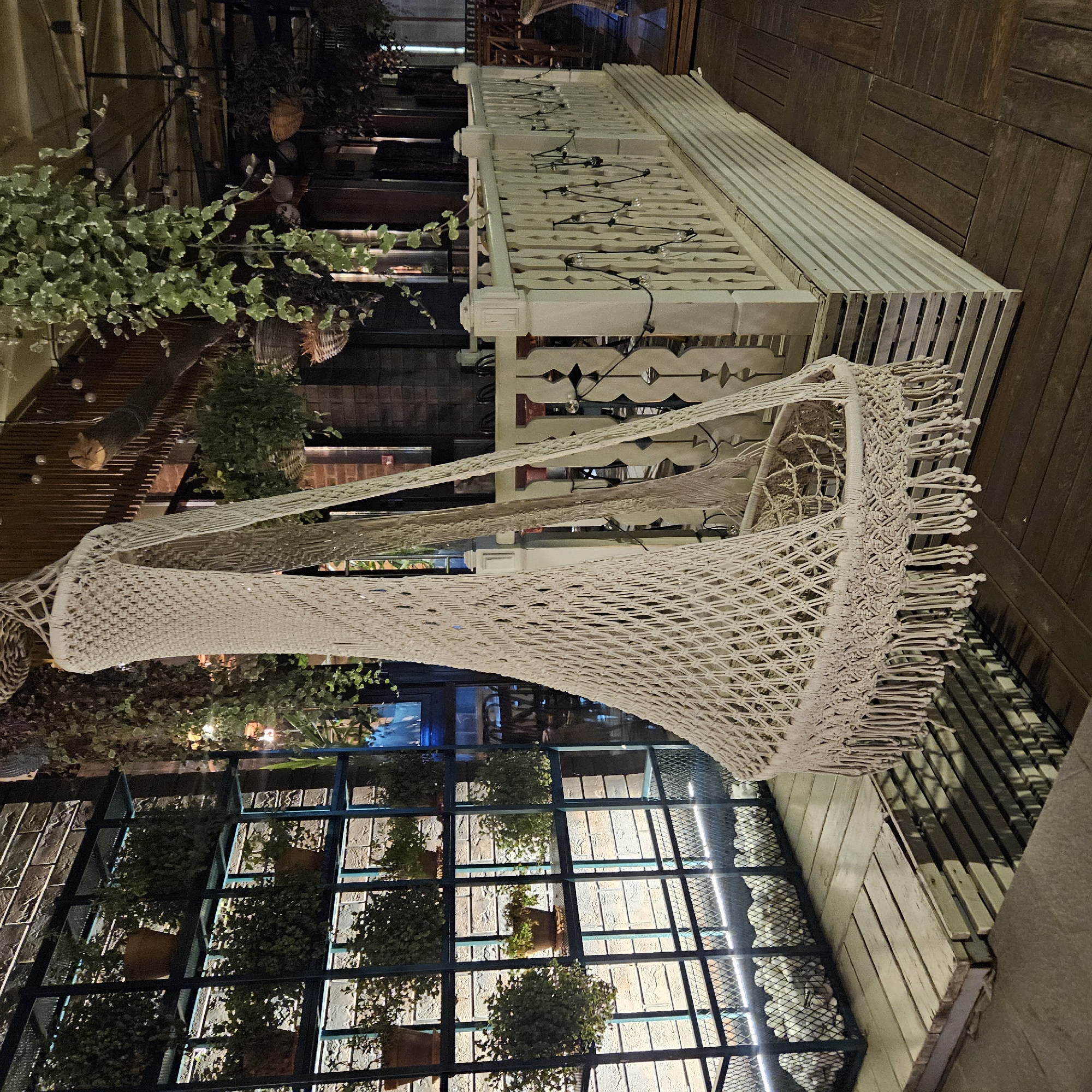 The image depicts an indoor scene, possibly a restaurant or cafe, with a macrame hammock chair as the central focus. The chair is white and intricately woven, hanging from the ceiling. The background reveals a brick wall, shelves filled with potted plants, and a wooden deck with a white railing adorned with string lights. Various hanging plants are visible, contributing to a natural and bohemian ambiance. There are no people or animals present in the image. The lighting appears to be dim, with artificial lights casting shadows on the wooden surfaces. The scene does not provide any specific visual cues that would indicate a location in Chelyabinsk, Russia.