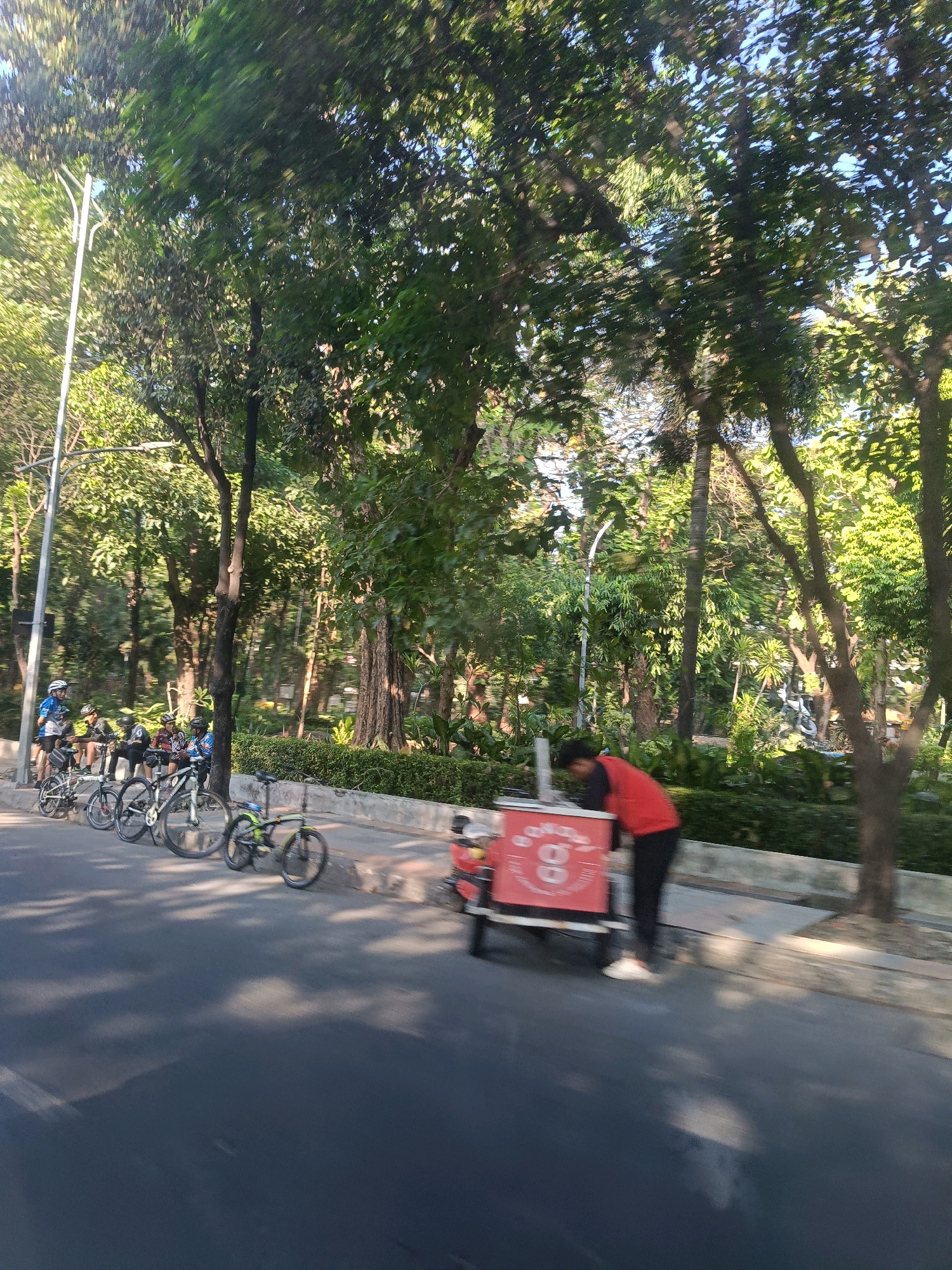 The image depicts a street scene in Surabaya, Indonesia, likely during daytime. In the foreground, a person wearing a red jacket and black pants is attending to a mobile food cart, which is predominantly red with white lettering and a circular logo. Several bicycles are parked along the sidewalk to the left, with a group of individuals wearing cycling gear standing near them. The background is filled with lush greenery, suggesting a park or tree-lined avenue. The road surface is asphalt, and sunlight casts shadows on it. The overall impression is a casual outdoor activity in an urban setting with abundant vegetation.