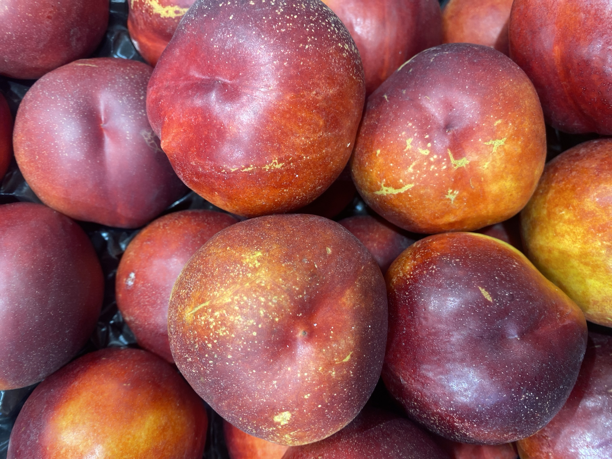 This image is a close-up, overhead shot of a pile of ripe nectarines at a market stall in Bel Air, Spain. The nectarines are primarily deep red with streaks of orange and yellow, and some are speckled with small white dots. The fruits are tightly packed together, suggesting they are for sale. The background is dark and indistinct, providing no further environmental details.