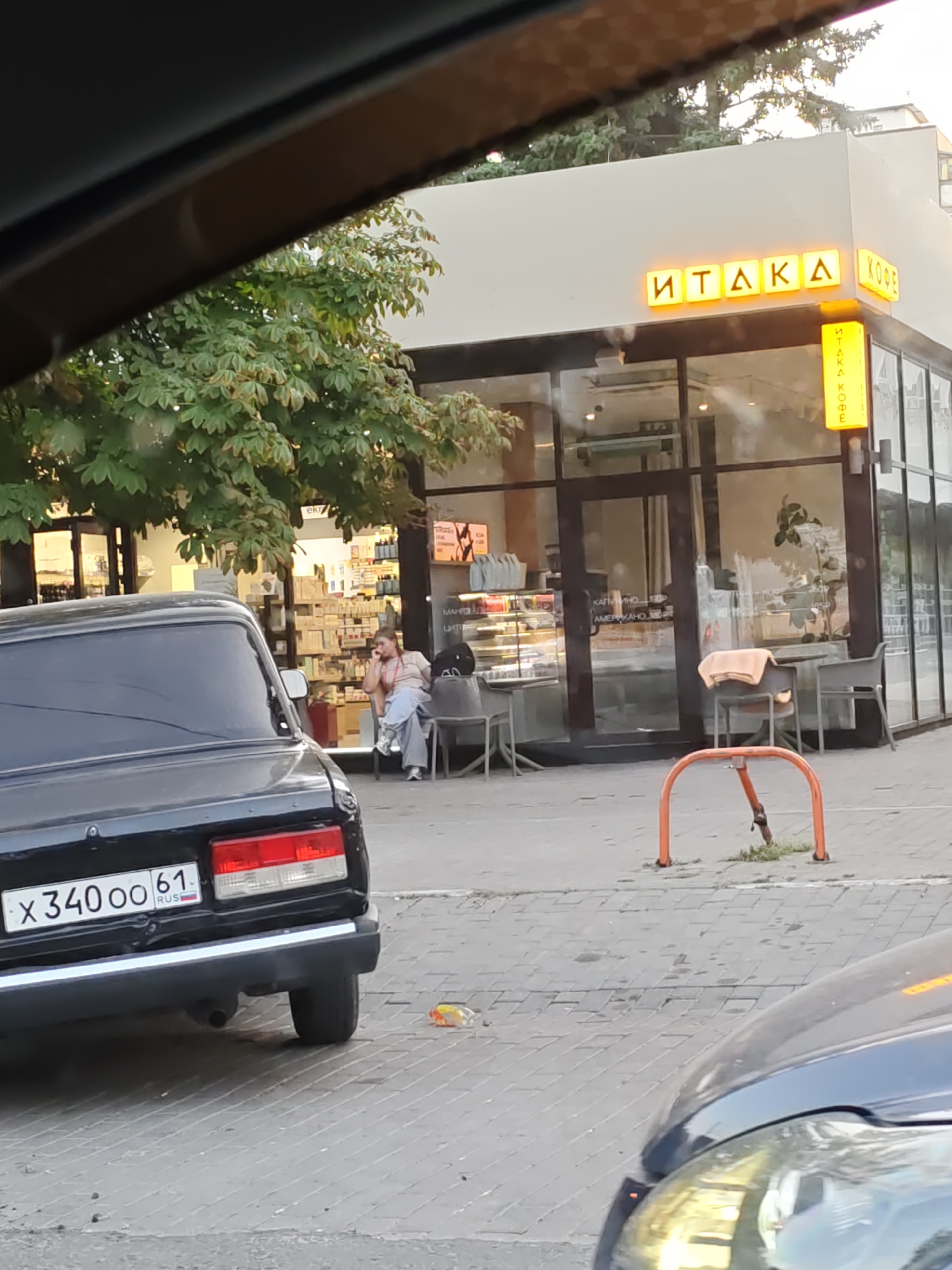The image shows a street scene in Rostov-on-Don, Russia. In the foreground, the rear of a black car with a license plate reading "X 340 OO 61 RUS" is visible. The car is parked on a paved surface. Behind the car, there is a storefront with a large glass window. The storefront has a sign that reads "ITAKA KOФE" in illuminated yellow letters on the facade, and a vertical sign with the same text. Inside the cafe, pastries and other items are displayed on shelves. Two people are seated outside the cafe on chairs. One person, appearing to be a woman wearing a light-colored top and blue pants, is sitting on a chair and holding a phone to her ear. Another person, possibly a man, is seated behind her. A table with a blanket draped over it is also visible outside the cafe. In the background, trees with green foliage are present, and there are buildings visible beyond the cafe. An orange bicycle rack is in the middle ground.