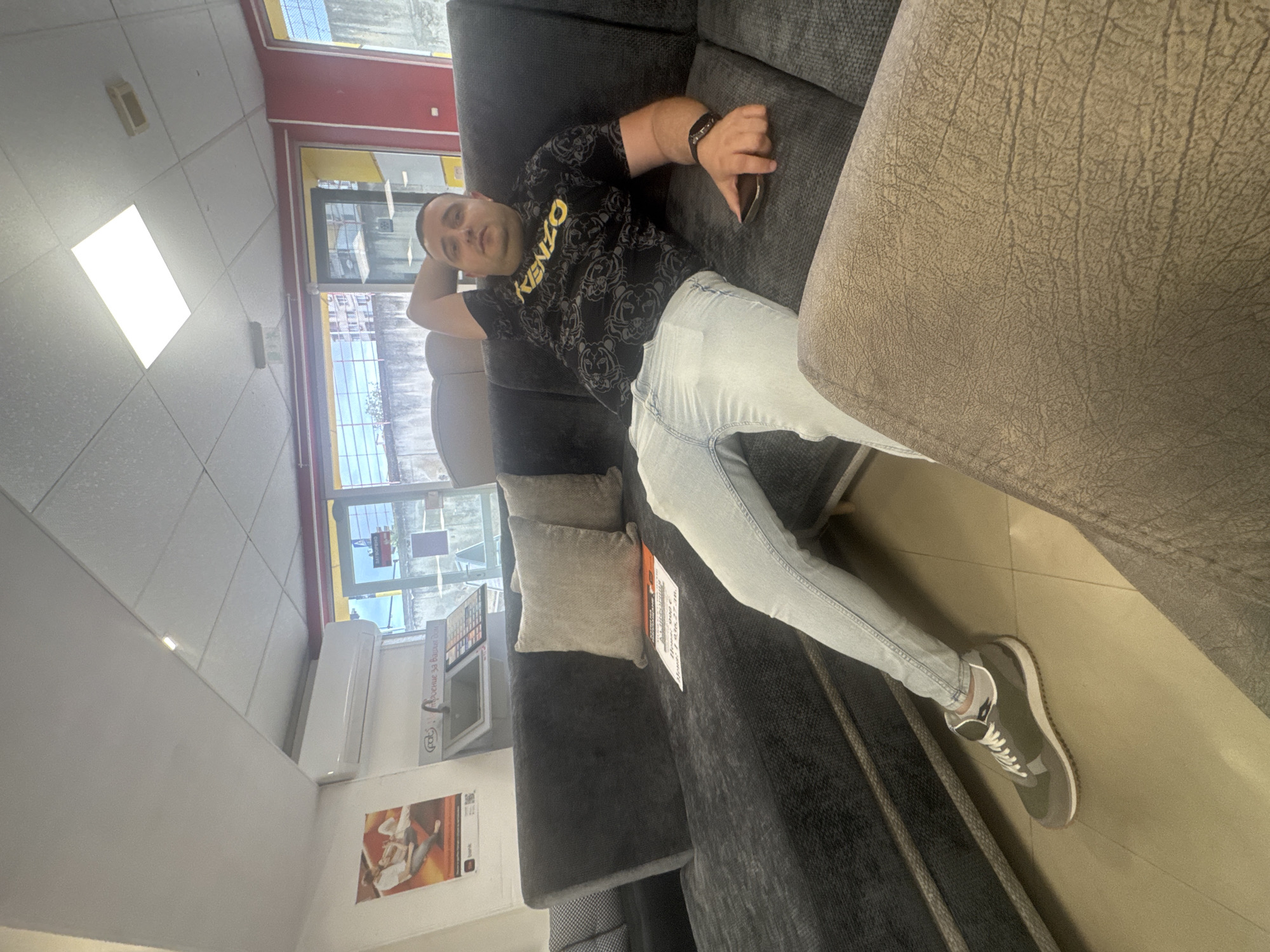 This image depicts an indoor scene, likely a furniture showroom or store in Shumen, Bulgaria. An adult male is the primary subject, seen relaxing on a dark grey upholstered sofa. He is leaning back with his left arm behind his head, appearing calm. He has short hair or is bald and is dressed in a black t-shirt featuring "KENZO" in yellow text, light blue jeans, and olive green and grey sneakers with white laces. He holds a dark-colored smartphone in his right hand and wears a dark bracelet on his right wrist.
The sofa is adorned with two light-colored cushions, and an adjacent section of a sofa in a lighter, textured brownish-grey fabric is visible to the right. The room features a tiled floor and ceiling, illuminated by bright rectangular overhead lights. A large window provides a view of an outdoor urban environment with buildings and possibly some greenery, indicating it is daytime. A white air conditioning unit is mounted on the wall below the window.
Notable text includes "KENZO" on the man's t-shirt. Near the window, a sign or screen displays a "POLIS" logo and partially legible Bulgarian text, including "Добавки за Баня" (Additives for Bath) and "Душ" (Shower). A white tag attached to the sofa clearly states a price: "Цена: 999 лв." (Price: 999 BGN), along with "Срок: 5 лв." (possibly a term or warranty duration) and "Кредит?" (Credit?), reinforcing the retail furniture store setting.