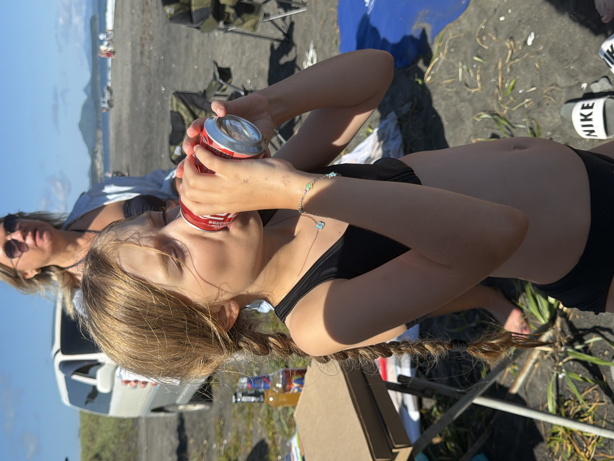 The image shows a group of people on a beach. A young girl is in the foreground, drinking from a red can. She has blonde hair braided and is wearing a black bikini top. Behind her, a woman wearing sunglasses and a light blue shirt is visible. The beach has dark sand and some vegetation. In the background, there are mountains and the ocean with waves. A white vehicle is also present on the beach. The scene appears to be set on a sunny day. The overall mood seems relaxed and casual. There are no prominent objects or text that provide specific details about the activity or event.
