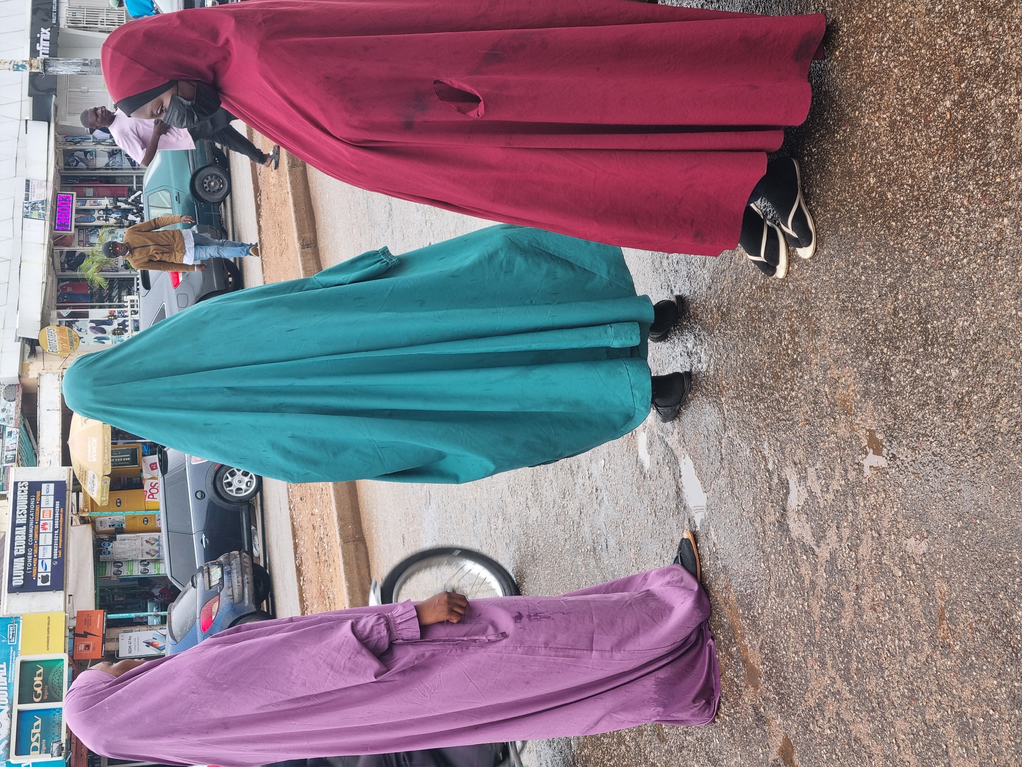 The image shows three women in colored abayas walking on a street in Jos, Nigeria. The abayas are burgundy, turquoise, and lilac. The woman in the burgundy abaya is wearing black and white sandals. The woman in the turquoise abaya is wearing black shoes. The woman in the lilac abaya is wearing brown shoes. The street is wet. Shops and vehicles are in the background. One shop has a sign that reads "OLUWA GLOBAL RESOURCES".
