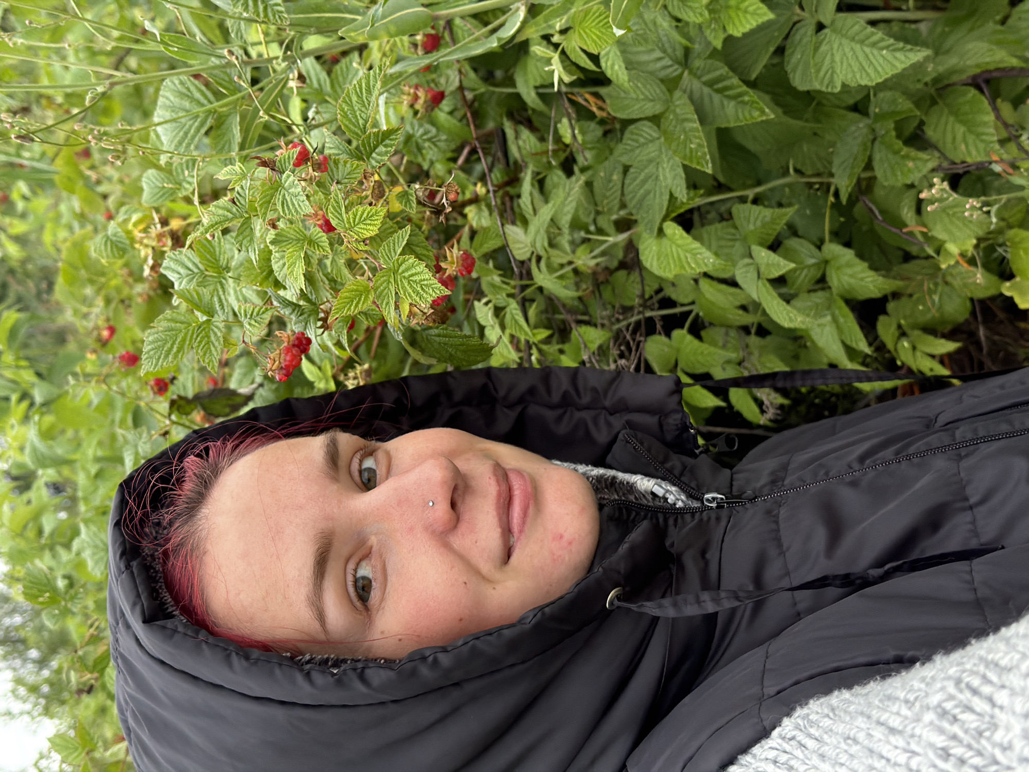 A young woman with red hair, wearing a black hooded jacket and a grey knitted sweater, is pictured from the chest up. She has a nose ring and is looking towards the camera with a slight smile. To her right, a dense patch of raspberry bushes with ripe red berries is visible. The background consists of lush green foliage, suggesting an outdoor, possibly natural environment. The image appears to have been taken during the daytime.