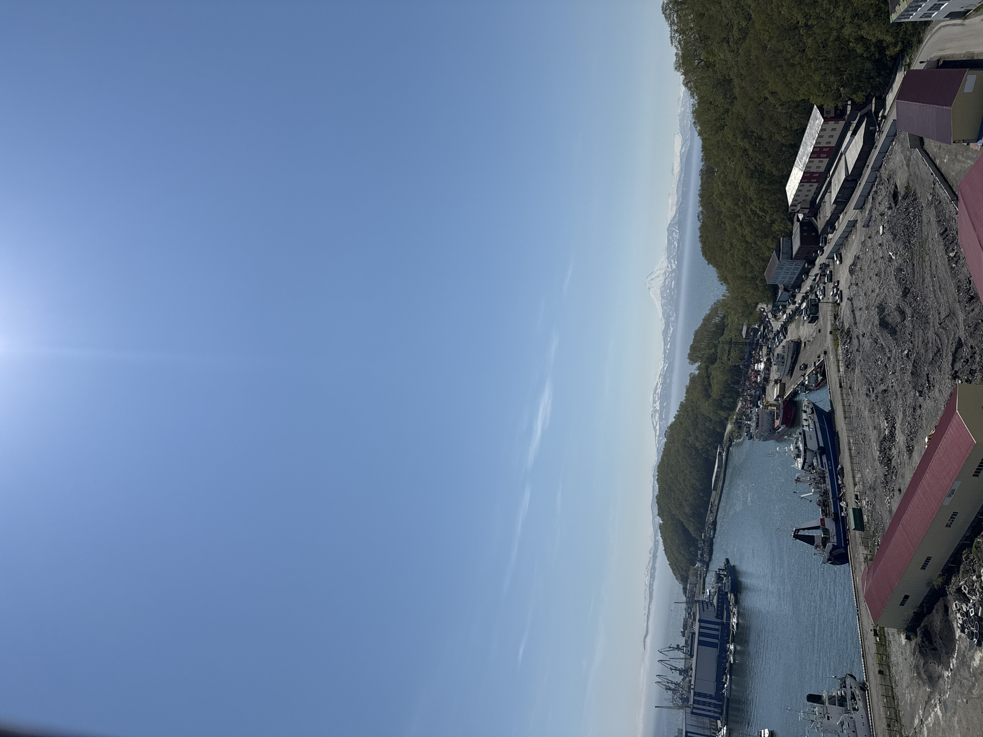 This is an aerial, high-angle, wide shot of a port and surrounding areas in Petropavlovsk-Kamchatsky, Russia, under a clear blue sky with faint wispy clouds. In the foreground, a large, elongated building with a red roof and yellow lower section is visible, with piles of dark soil and debris around it. To the left, another building with a red roof is partially visible.
In the middle ground, a harbor is filled with various vessels, including large fishing trawlers and smaller boats. Cranes and industrial structures line the port. Adjacent to the harbor, a densely wooded hillside rises, with a road winding through it, lined with parked vehicles and several buildings, including a long building with a red and white facade and another with a light blue facade. Further inland, snow-capped mountains form a dramatic backdrop, with a prominent, cone-shaped volcano dominating the distant skyline. The sea is visible between the port and the mountains. The overall impression is a bustling port town set against a rugged, mountainous natural landscape.