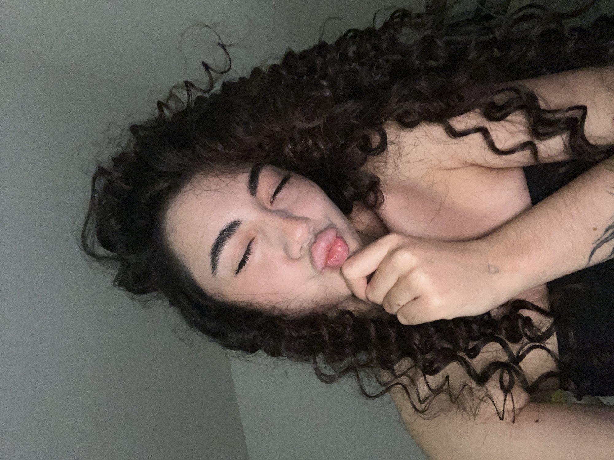 A young woman with long, curly dark hair is pictured from the shoulders up. Her eyes are closed and her lips are pursed in a kissing gesture, with her right hand touching her chin. She is wearing a dark top. The background is a plain, light-colored wall. The image appears to be a selfie taken indoors, with soft lighting. There is no indication of the location being in Chotěšov, Czechia.