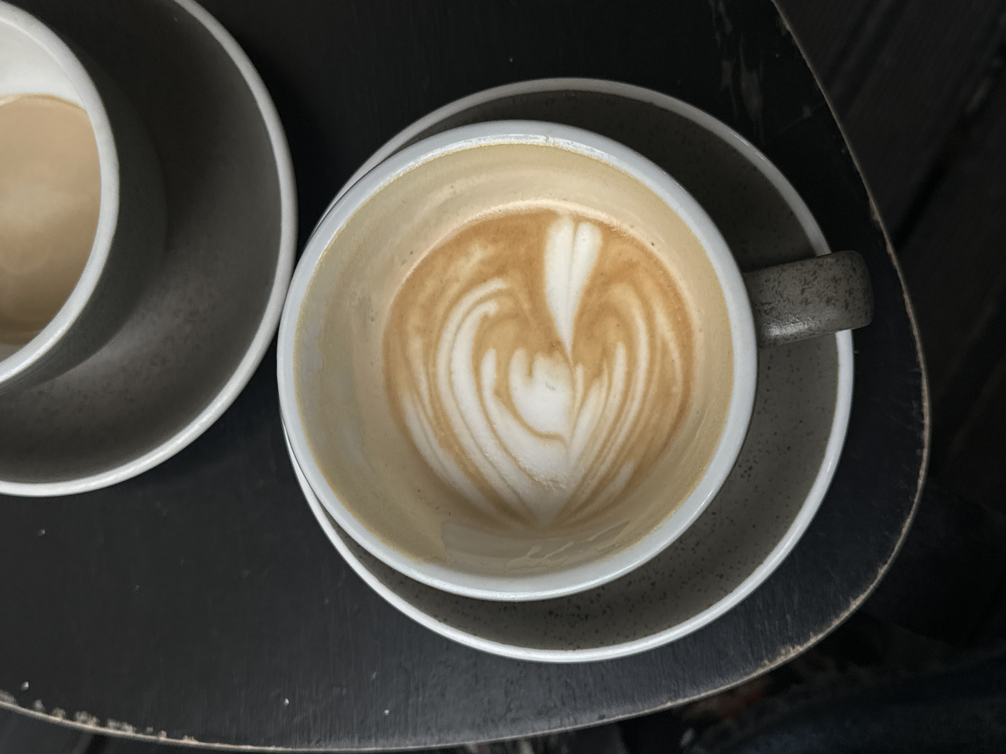 The image shows two cups of coffee on a dark, round table. The cup on the right is nearly empty, revealing latte art in the shape of a heart. The cup on the left contains coffee with milk. The table appears to be indoors, likely in a cafe. The location is presumed to be Paris, France.