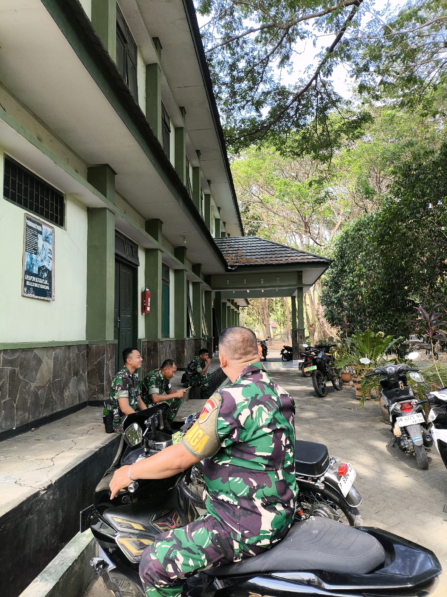 The image depicts an outdoor scene in front of a building, likely a military facility in Indonesia. The building has a green exterior with white trim and a tiled roof. In the foreground, a person in a camouflage uniform is seated on a black motorcycle, facing away from the camera. Their left hand rests on the handlebar, and a bracelet is visible on their wrist. To the left of the motorcycle, three other individuals in similar uniforms are seated on the ground, facing the building. One is looking at a mobile phone, another is looking towards the right, and the third is looking towards the left. Several motorcycles are parked in the background. The environment is characterized by trees and foliage, suggesting a tropical or subtropical location. A poster is affixed to the building's wall on the left.