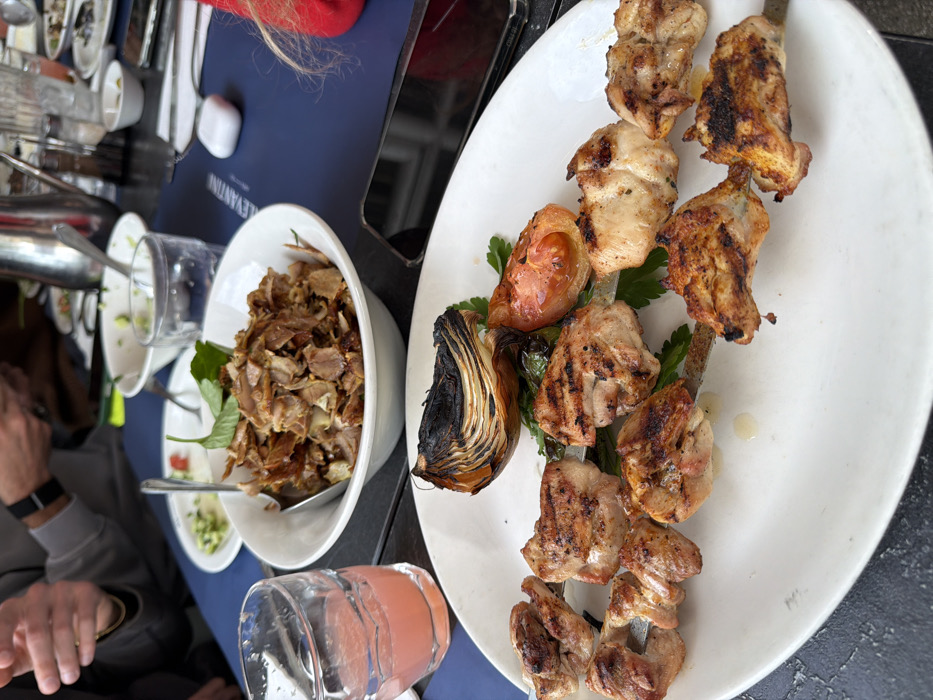 The image depicts a dining table scene at the Levantin restaurant in Tel Aviv, Israel.
In the immediate foreground, a white oval plate presents two metal skewers laden with grilled chicken pieces, characterized by golden-brown coloration and visible char marks. Accompanying the skewers are a charred, quartered onion, a grilled tomato half, and scattered green parsley sprigs. Adjacent to this plate, on the left, a textured clear glass holds a pink-hued beverage.
In the mid-ground, a white circular bowl contains a substantial portion of sliced, grilled meat, consistent with shawarma or similar preparations, garnished with green herbs. A silver spoon rests within this bowl.
The table surface is dark, with a blue placemat prominently featuring the text "LEVANTIN" in white lettering. Resting on this placemat are a black smartphone, placed face down, and a white wireless earbud charging case.
In the background, portions of two individuals are visible. One person's hands are seen, with a wrist wearing a watch, suggesting a male. This individual is wearing a grey long-sleeved garment. A small section of another person's red clothing is visible on the far right. The background also reveals additional dining accoutrements, including various small white bowls, glasses, a silver pitcher, and salt and pepper shakers, indicating an active restaurant environment and ongoing meal service.