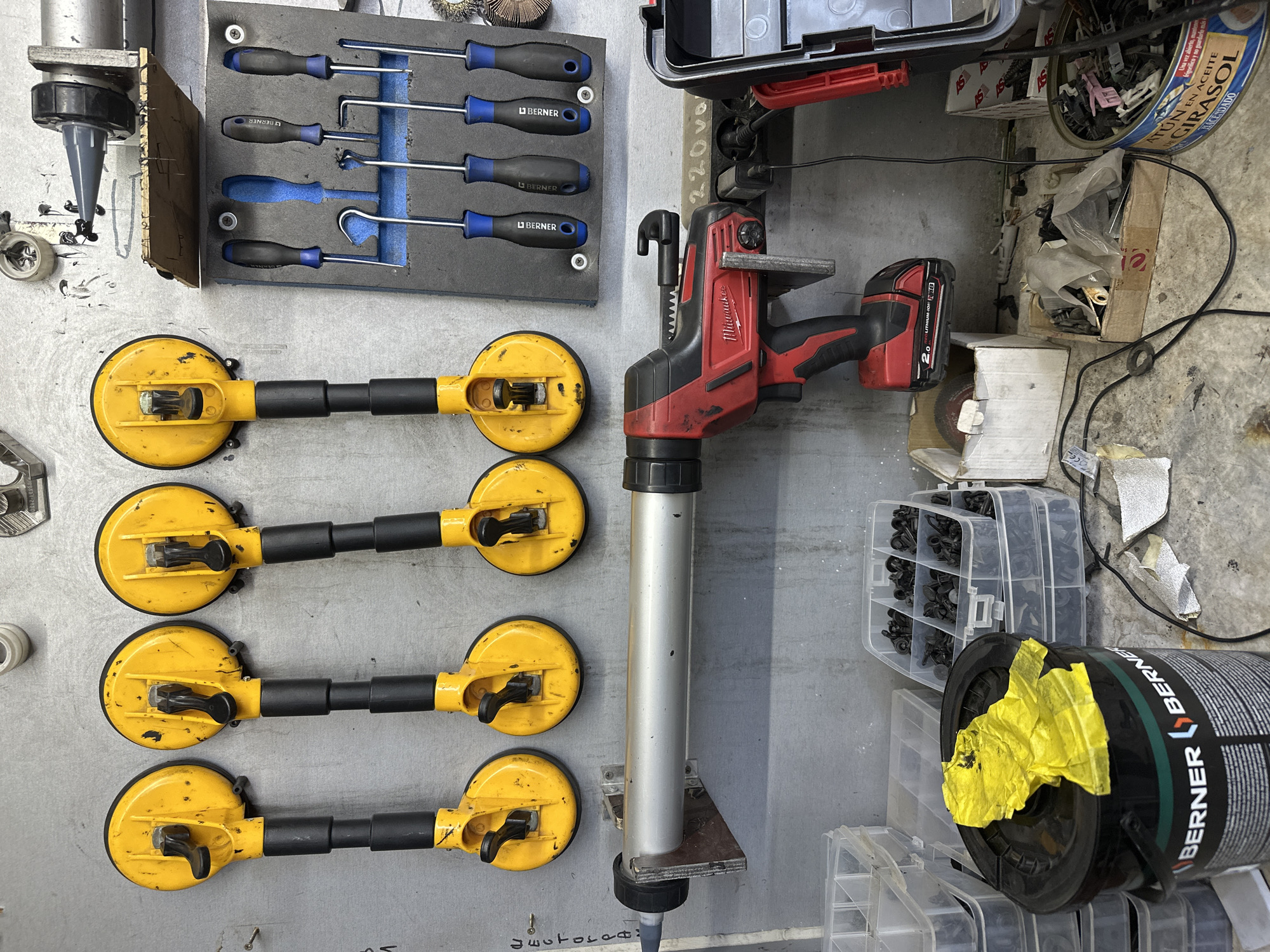 The image depicts a workshop or garage setting with a tool-laden wall. Mounted on the wall are four yellow suction cup lifters, each with a black handle. Below them, a silver caulking gun is attached to a bracket. To the right of the suction cup lifters, a foam board holds an assortment of screwdrivers with blue and black handles, as well as some L-shaped tools. A red and black power drill is positioned below the caulking gun, with a power cord plugged into an outlet. In the lower portion of the image, various storage containers filled with small black parts are visible, along with a large black bucket labeled "BERNER." The overall environment appears to be organized with tools and supplies for various tasks.