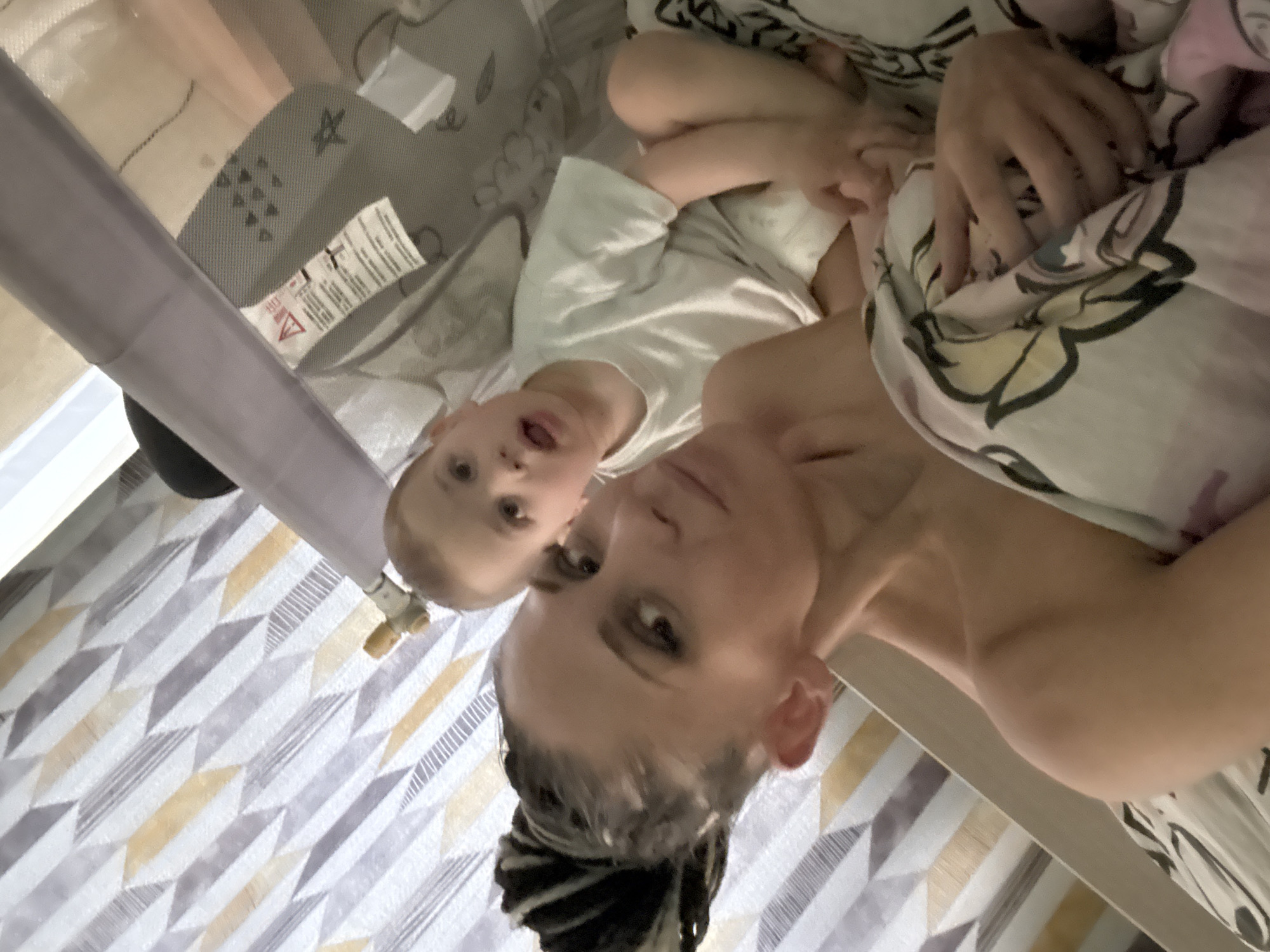 The image captures a close-up, candid moment of a woman and a young child, likely a mother and her son, relaxing together in an indoor setting. The photo is taken from a high angle, looking down at the subjects who are lying on their backs, giving the impression of an intimate, overhead perspective.
The woman is in the foreground, partially visible, looking directly towards the camera. She has dark hair styled in dreadlocks or braids pulled back, and her bare shoulder is visible. Her expression is calm and neutral. Behind and slightly above her, a young child, appearing to be a boy, is also looking towards the camera with his mouth slightly open. He has light-colored hair and is wearing a white t-shirt.
They appear to be on a bed or inside a large playpen or crib. The mattress is covered with a sheet featuring a geometric pattern of white, gray, and yellow shapes. To the left and top of the frame, a gray mesh side of a playpen or crib is visible, along with a gray structural bar. A white label with a red triangular warning symbol (an exclamation mark) and some unreadable text is attached to the mesh, likely safety information. Parts of a light-colored blanket or duvet with a muted, patterned design (possibly floral or abstract) are draped over the woman and child. A small, golden, valve-like object is also visible attached to a vertical element in the bottom-left area of the frame.
The scene suggests a relaxed moment of connection, possibly during playtime or rest, within a home environment like a bedroom or nursery. The lighting is soft and even, offering no specific cues for the time of day.