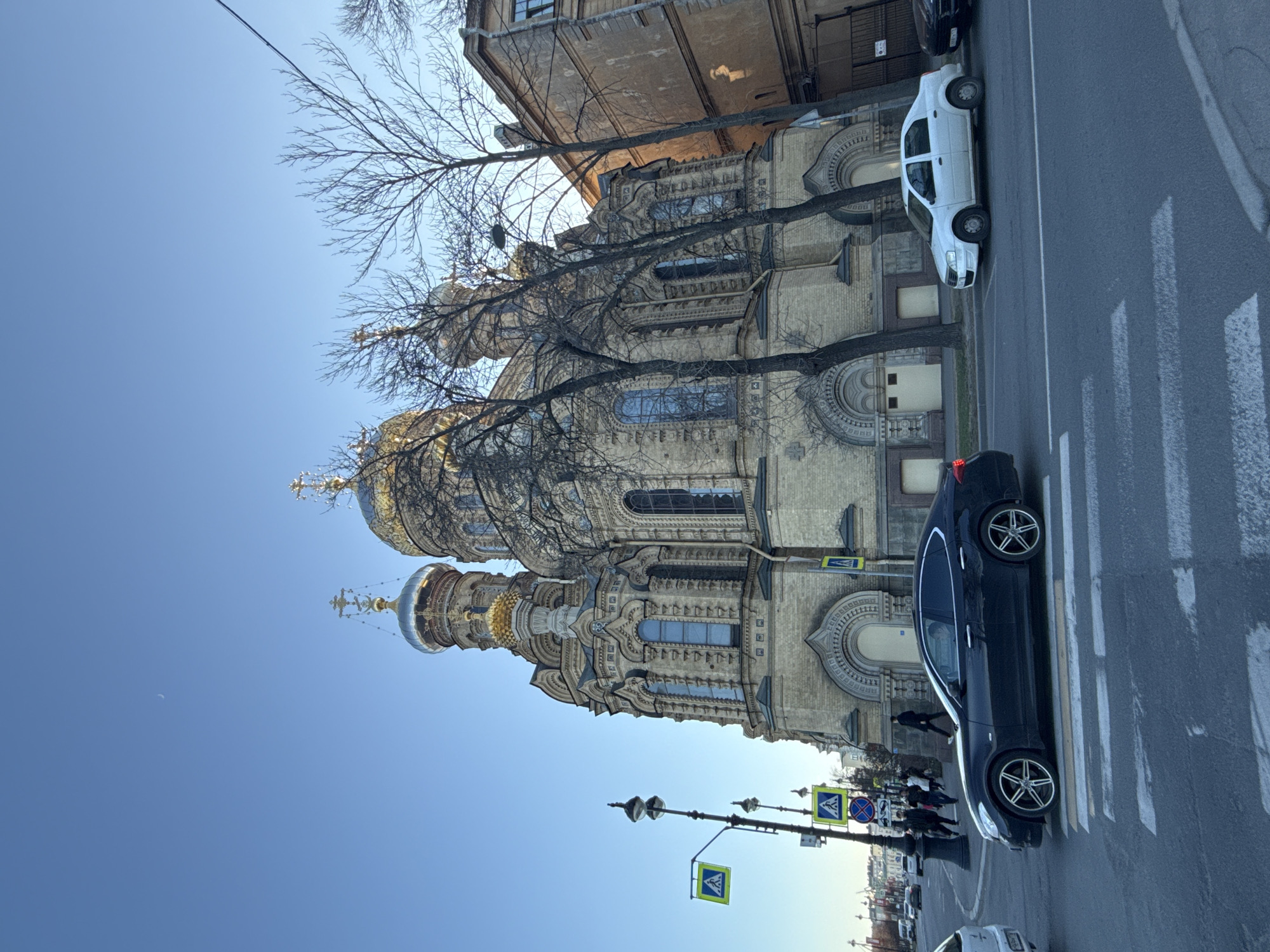 This media file presents a bright, sunny street scene in Saint Petersburg, Russia, featuring a grand and ornately decorated church. The image, which appears to be rotated 90 degrees clockwise, captures the multi-domed edifice in a clear blue sky.
The main subject is a magnificent church, characterized by its intricate architecture, featuring multiple domes—some golden, some with a silver or reflective finish—each topped with an Orthodox cross. The church facade is richly detailed with elaborate carvings, decorative columns, and arched windows. Bare deciduous trees stand in front of the building, their branches creating a dark, intricate pattern against the lighter tones of the church and sky, suggesting late winter or early spring.
The sky is a clear, vibrant blue, occupying a significant portion of the upper left side of the image (when viewed upright). A subtle crescent moon is visible against the blue, indicating either early morning or late afternoon.
At street level, an asphalt road with clear white zebra crossing markings is prominent. A dark-colored sedan with distinctive multi-spoke alloy wheels is positioned near the crosswalk. Further along the street, a white van is parked on the side. Several indistinct figures of people can be seen walking on the sidewalk in the middle distance. Street furniture includes a lamppost and a pole with various traffic signs, notably a blue square pedestrian crossing sign and a circular "no parking" or "no stopping" sign.
The overall scene depicts a typical urban environment with pedestrians and vehicles, set against the striking backdrop of historical Russian architecture, under clear weather conditions.