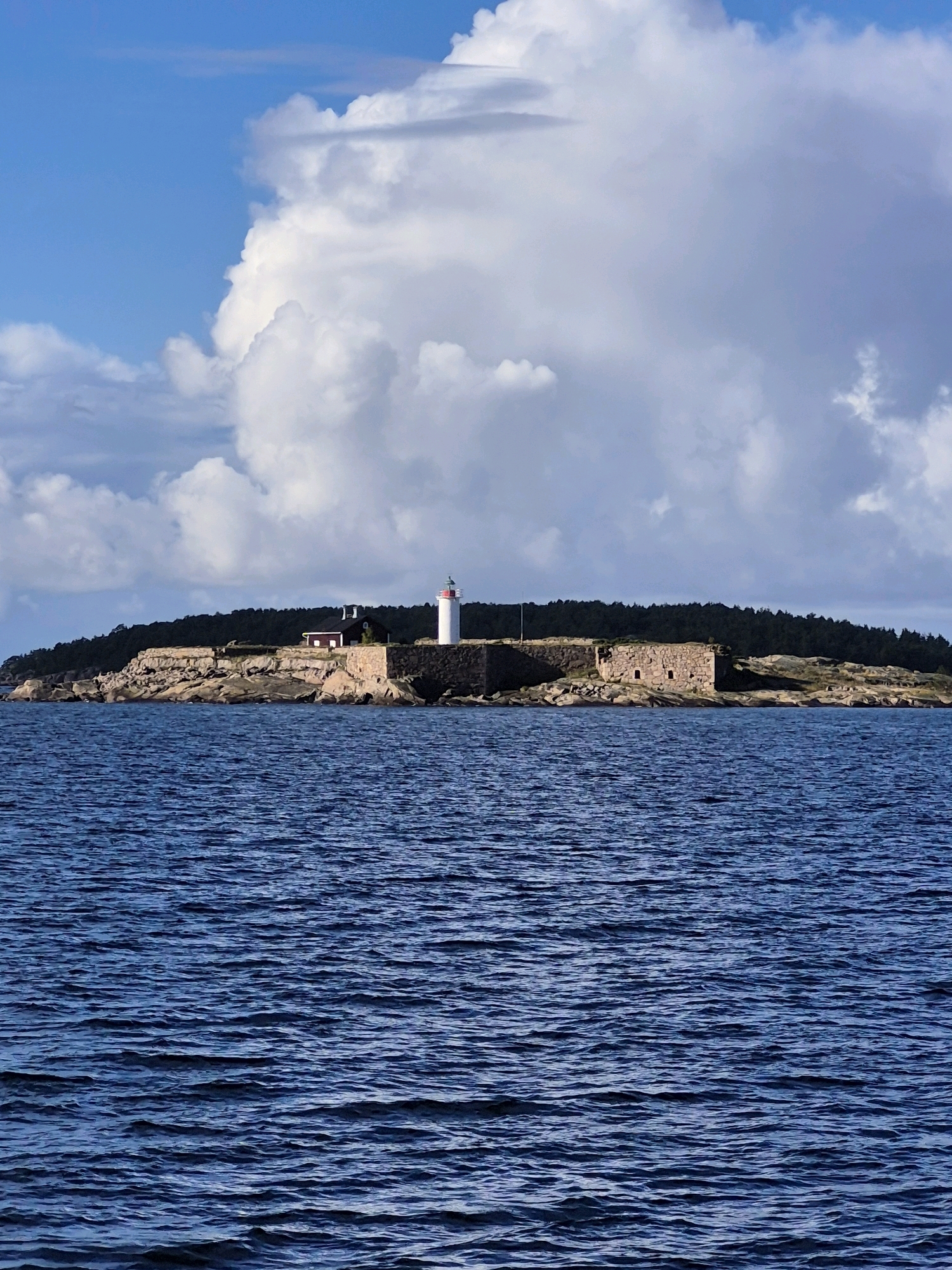The image shows a lighthouse and fort ruins on an island in the sea, likely near Hanko, Finland. The sea is a deep blue with small waves, and the sky is partially cloudy with large white cumulus clouds dominating the upper portion. The island is covered with rocks and vegetation, with a dark green forest line in the background. The fort ruins are made of stone and appear old. A white lighthouse with a red top stands near a small red building with a dark roof. The scene is calm and natural, with no people visible.