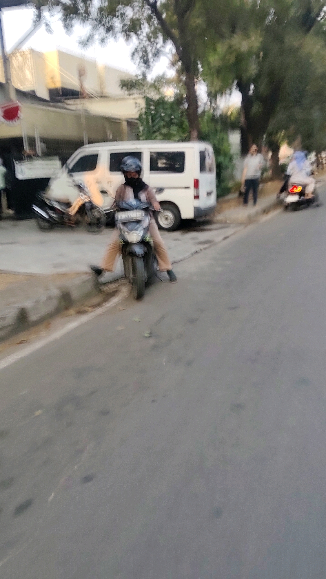 In the city of Mekarmukti, Indonesia, a blurred image captures a street scene. A person, wearing a helmet and light brown pants, sits on a dark-colored motorcycle that is parked facing forward, with its front wheel angled slightly to the right. The motorcycle's license plate, bearing the inscription "R 2921 ERB," is partially visible. Behind the motorcycle, a white van is parked, with its side door slightly ajar. To the right of the van, two individuals are visible in the distance: one standing and the other on a motorcycle, both out of focus. The street itself is paved, with a curb and some foliage bordering the roadway. The overall lighting suggests it is either early morning or late afternoon.