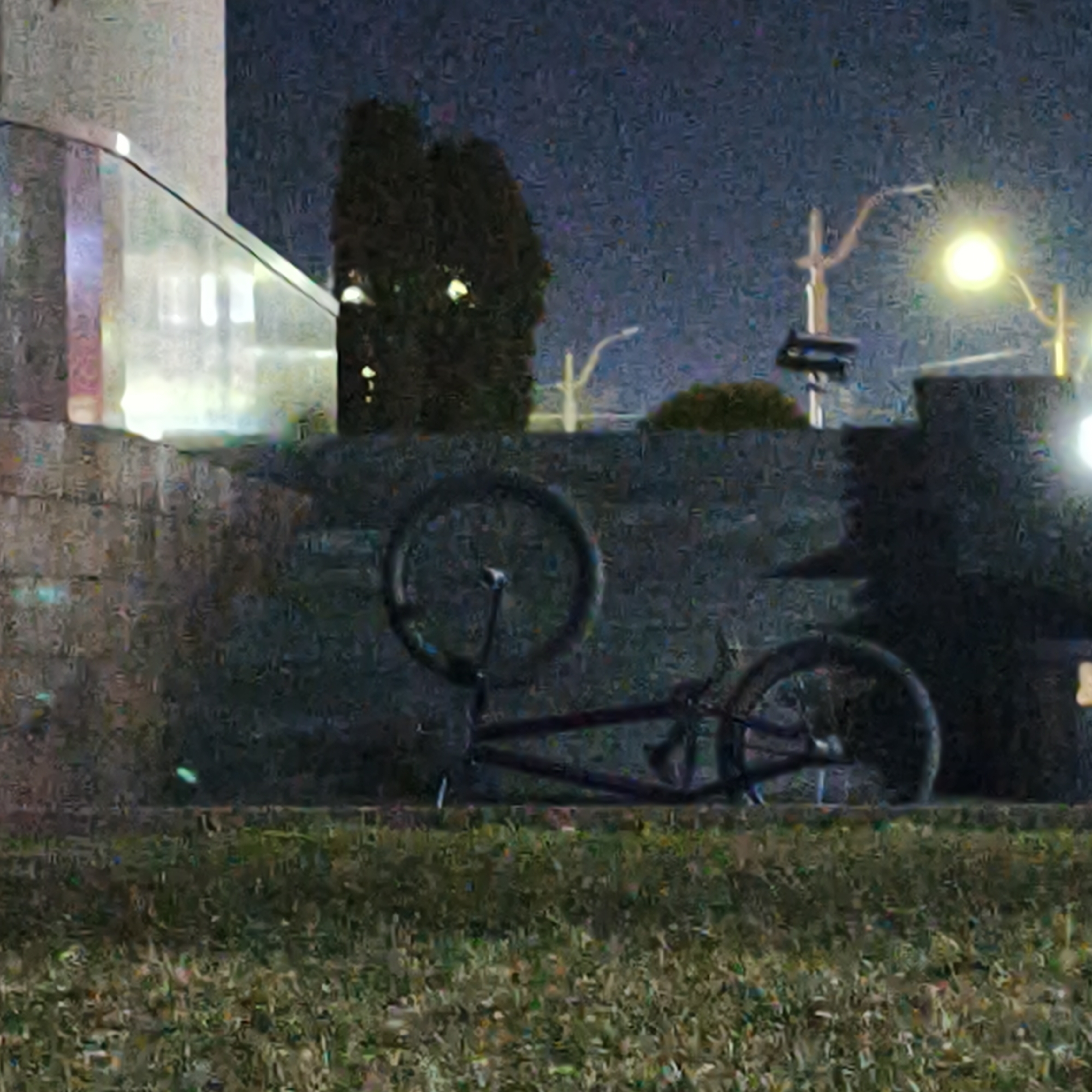 The scene is a nighttime outdoor view located in Hrodna, Belarus. In the immediate foreground, the ground is covered with natural green-brown grass. A dark-colored, BMX-style bicycle lies horizontally on its left side on the grass, oriented with its front wheel to the right and rear wheel to the left. Behind the bicycle, a dark, textured wall or embankment, likely constructed of stone or concrete, spans the midground. Above and beyond this wall, to the left, a modern architectural structure is partially visible, featuring a light-toned vertical element and a curved glass railing reflecting internal light sources. To the right, a prominent street light emits a bright yellow-white illumination, positioned above and beside a dark, dense bush or tree. The sky overhead is dark. No individuals or active subjects are present. The image exhibits high visual noise and low resolution.