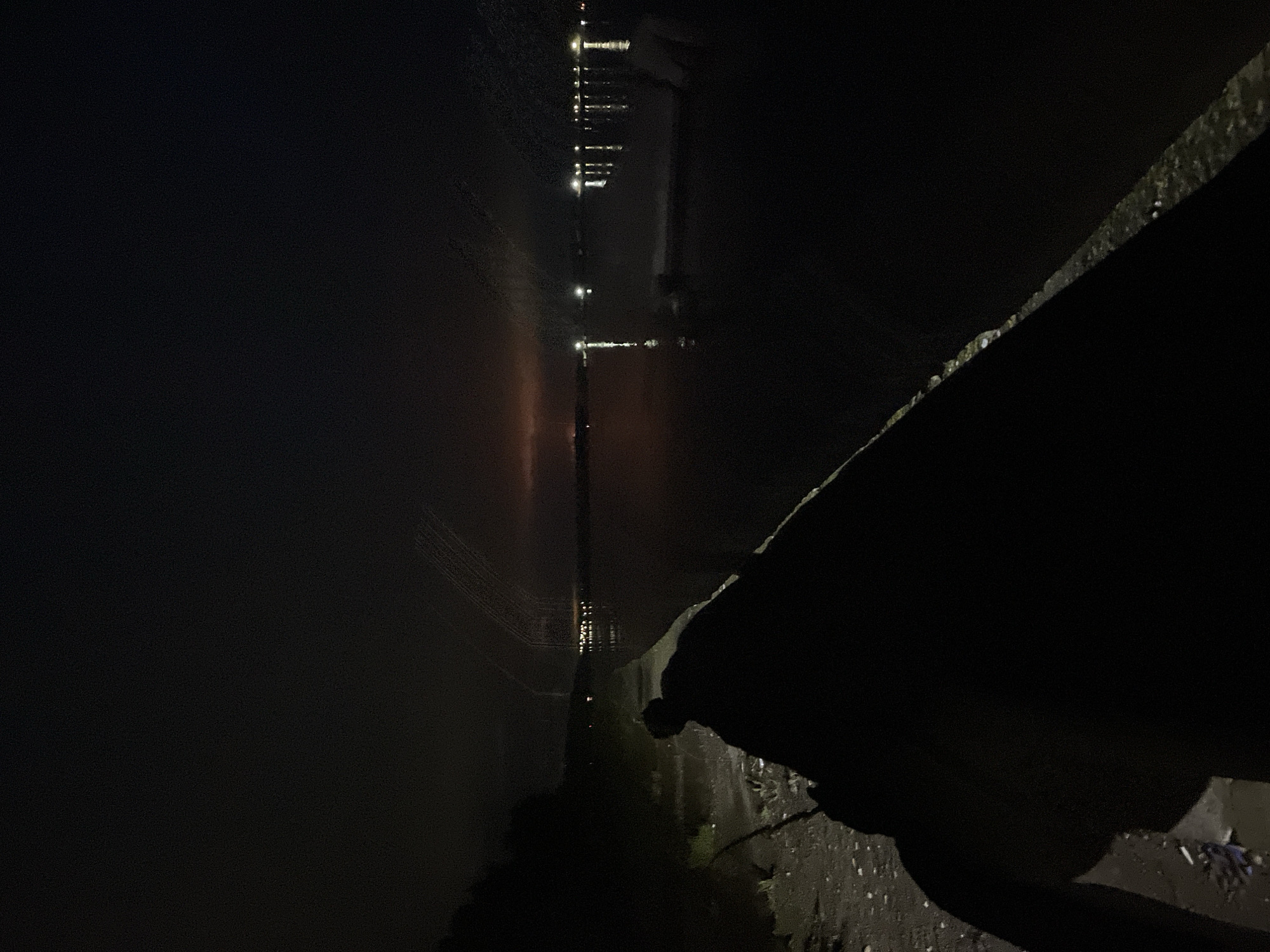 This nighttime image, captured in Chaikovsky, Russia, depicts a dark, expansive scene by a body of water, likely a river or lake.
In the foreground, a heavily silhouetted figure, presumed to be a person, is positioned in the lower-right quadrant, partially obscured by a concrete embankment. A rounded form at the top of the silhouette suggests a head or cap. A thin, dark rod, consistent with a fishing rod, extends from the lower-left corner of the frame, diagonally towards the center, its tip barely visible near the water's edge. The immediate ground to the left of the embankment is a dark, granular surface, appearing as a dirt or gravel path, with scattered small, light-colored specks, likely pebbles or debris. A small piece of blue material is visible on the ground in the bottom-left. Sparse, dark vegetation occupies the far left edge of the scene.
A concrete or stone embankment forms a distinct diagonal line from the lower-middle towards the mid-right, separating the land from the water. Its surface exhibits irregular lighter patches. The body of water, occupying the central and right portions of the image, is dark and still, reflecting distant light sources.
In the background, across the water, multiple points of artificial light are visible, casting elongated vertical reflections on the water's surface. These lights indicate a distant settlement or industrial area. The sky above the distant shore is dark, with a noticeable reddish-orange glow on the horizon towards the left, suggesting light pollution or a specific luminous source. Faint, parallel, and somewhat curved linear streaks are discernible in the upper sky, above the distant lights, appearing as atmospheric phenomena.