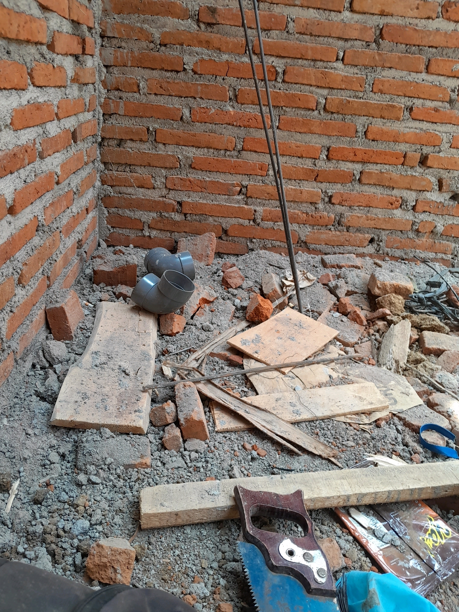 The image depicts a construction site in Karangduren, Indonesia. The scene is an interior corner of a building under construction, evidenced by the exposed red brick walls and rough mortar. The ground is covered in debris, including broken bricks, rubble, and pieces of wood. Several construction materials are scattered around: two grey PVC pipe elbows are resting on a piece of wood, two vertical metal rods are leaning against the wall, and a hand saw with a blue blade and a brown handle lies on the ground near some packaging materials. A blue strap is also visible amongst the debris. The lighting suggests it is daytime, and the overall atmosphere is one of active construction work.