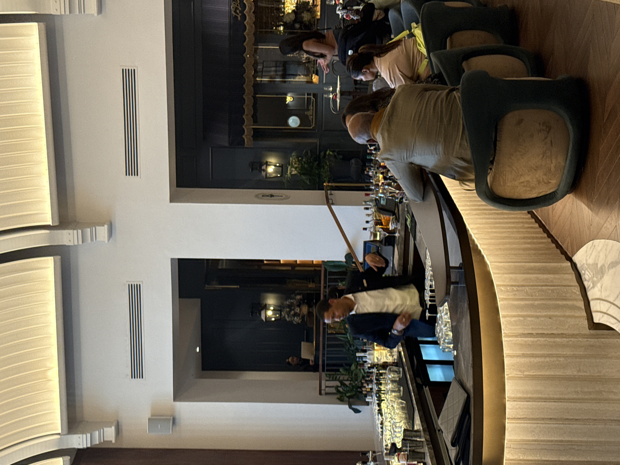 This media file captures an interior view of a stylish, upscale bar or restaurant, likely during the evening in Cape Town, South Africa.
The scene features a long, curved bar counter with strip lighting along its edge, creating a warm glow. Behind the bar, numerous bottles of alcohol and various types of glassware, including wine glasses and tumblers, are neatly arranged. A bartender in a white shirt and dark jacket is visible behind the counter, appearing to be focused on tasks.
Several patrons are present. On the right, two individuals are leaning on the bar, one with a bald head wearing a light-colored shirt, and another with dark hair, both viewed from behind. Next to them, a person in a light-colored top with a yellow bag is seated on one of the dark green bar stools, which feature tan-colored seats.
The decor includes architectural elements such as a curved, ribbed white wall feature on the left with indirect backlighting. Rectangular air vents with vertical slats are embedded in the white walls. In the background, beyond the bar area, dark-framed architectural elements suggest another section of the establishment, possibly with an awning and lantern-style lights, giving a sophisticated feel. Potted plants add a touch of greenery to the dimly lit space. The flooring is made of wooden parquet.
The overall ambiance is relaxed and inviting, typical of a lounge or bar setting. There is no clearly legible text visible in the image.