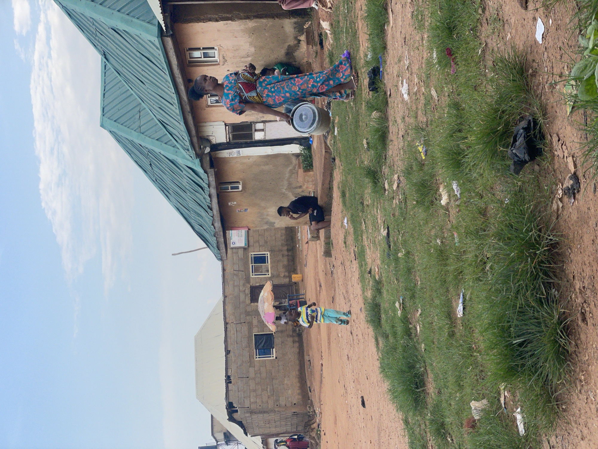 In Kurunduma, Nigeria, a young child in a striped t-shirt and blue pants runs across a dirt path. Nearby, a woman in a vibrant, floral-patterned dress walks with a bucket in her right hand. Behind them, a building with a green, corrugated metal roof stands under a cloudy sky. Other people are visible in the background, some sitting outside homes and others tending to an umbrella shelter. The environment appears somewhat unkempt, with grass patches interspersed with scattered debris.