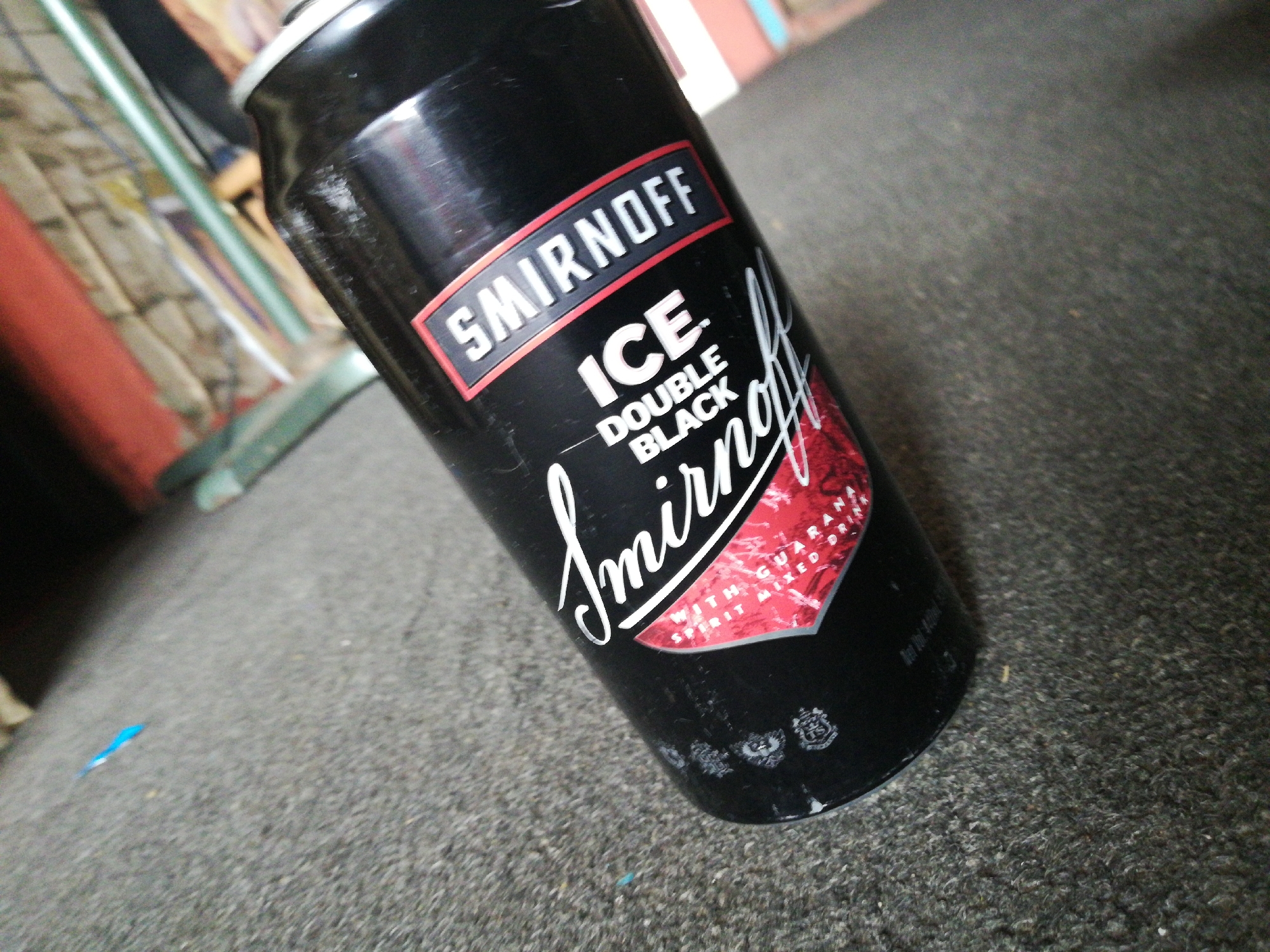 A black can of Smirnoff Ice Double Black is sitting on a rough, grey surface, possibly carpet or concrete. The can is angled slightly, with the Smirnoff logo clearly visible. The background is blurred, showing a chair with green legs and a partially visible curtain. The location is Unknown, Unknown.