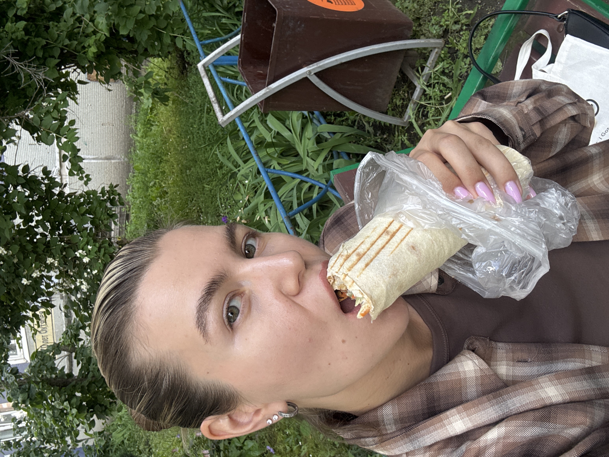 A young woman is captured in a selfie outdoors, in what appears to be a courtyard or park area. She is holding a shawarma wrapped in foil and plastic wrap, and is in the process of taking a bite. The woman has her hair pulled back and is wearing a plaid shirt over a brown t-shirt. Her left ear has two small hoop earrings. The background shows greenery, a building facade with a sign in Russian text, a blue metal fence, and a brown trash receptacle. The lighting suggests it is daytime.