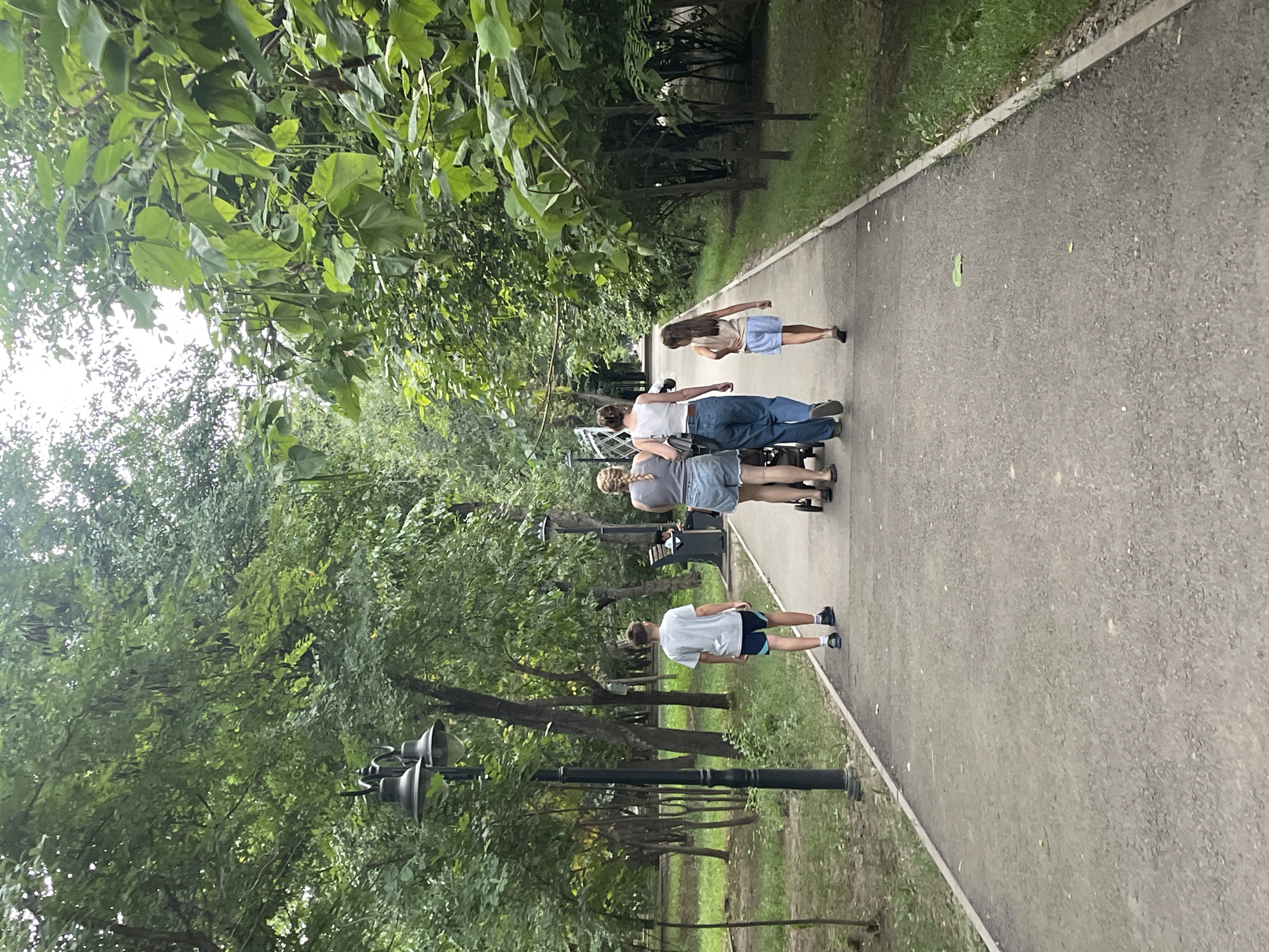 The image shows a paved pathway in a park, lined with trees and grass. The pathway is curved, leading into the distance. There are a few lampposts on the left side of the path. In the foreground, a young boy in a grey t-shirt and blue shorts walks away from the camera. Further down the path, a woman in a white tank top and blue jeans pushes a stroller, accompanied by another woman in a grey tank top and denim shorts. A young girl in a tan top and blue shorts walks behind them. The foliage is lush and green, suggesting a warm season. The overall atmosphere is peaceful and leisurely. The scene is located in Almaty, Kazakhstan, likely within a public park such as Park of 28 Panfilov Guardsmen or Central Park.