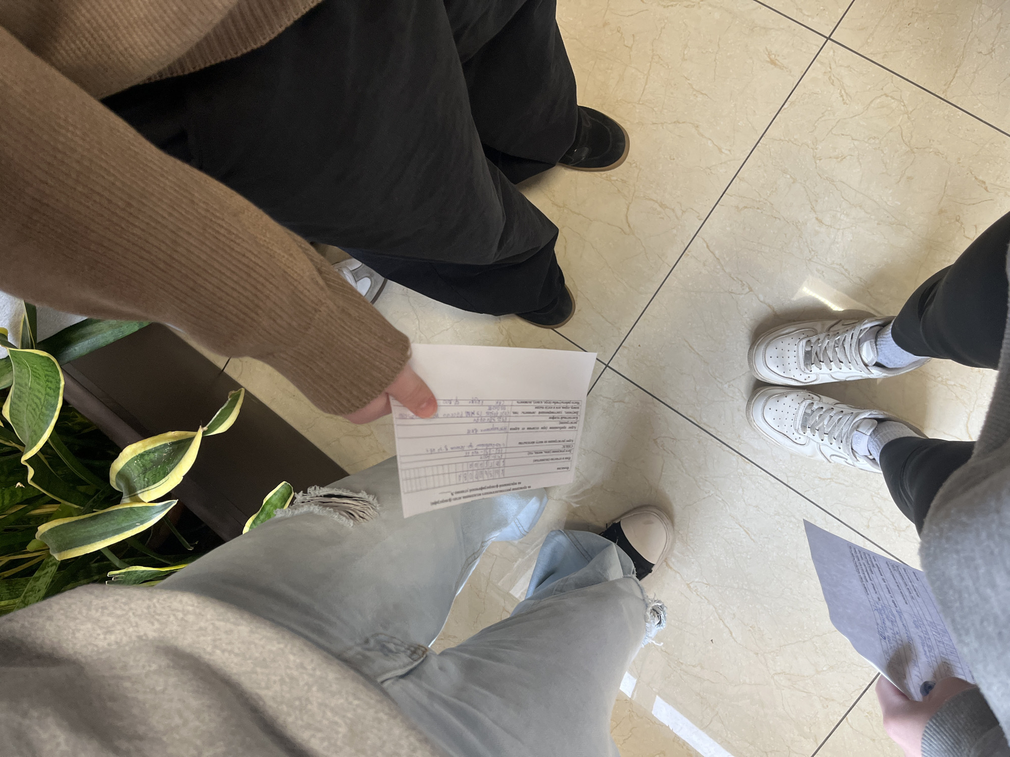The image shows a view of a tiled floor with three people standing on it, each holding a piece of paper. The floor tiles are light beige with darker grout lines. The person on the left is wearing light blue ripped jeans, a gray sweater, and black and white sneakers. They are holding a piece of paper with visible text in their hand. The person in the top center is wearing a tan sweater and dark pants with black shoes. The person on the right is wearing white sneakers, dark pants, and a gray sweater. They are also holding a piece of paper. There is a green potted plant visible on the left side of the image. It is not possible to determine the location based on the image.