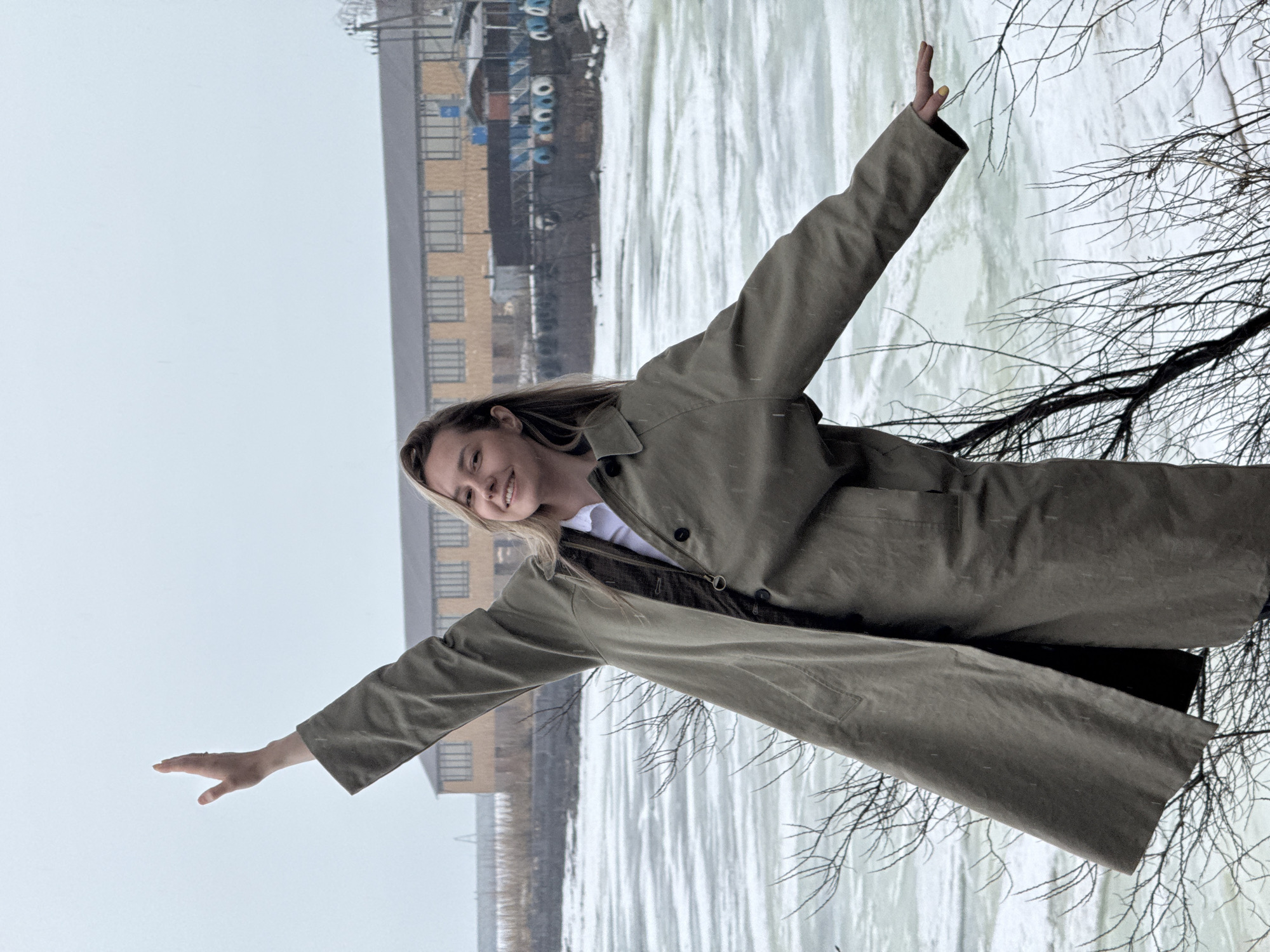 A woman stands in front of a body of water, likely a river or a bay, with a building in the background, seemingly in Chaikovsky, Russia. The body of water has a partially frozen surface, creating a mixture of ice and open water.
The woman is wearing a long, dark green or olive-colored coat over a white shirt and dark pants. She has her arms outstretched, as if embracing the scene or balancing herself.
The background features a multi-story building with a yellowish brick facade. Beyond the building, there's a structure with blue cylindrical objects attached, possibly part of an industrial or port facility.
Bare tree branches are visible on the right side of the image, suggesting a winter or early spring season. The sky is overcast, contributing to the muted color palette of the scene. There is evidence of snowfall in the image.