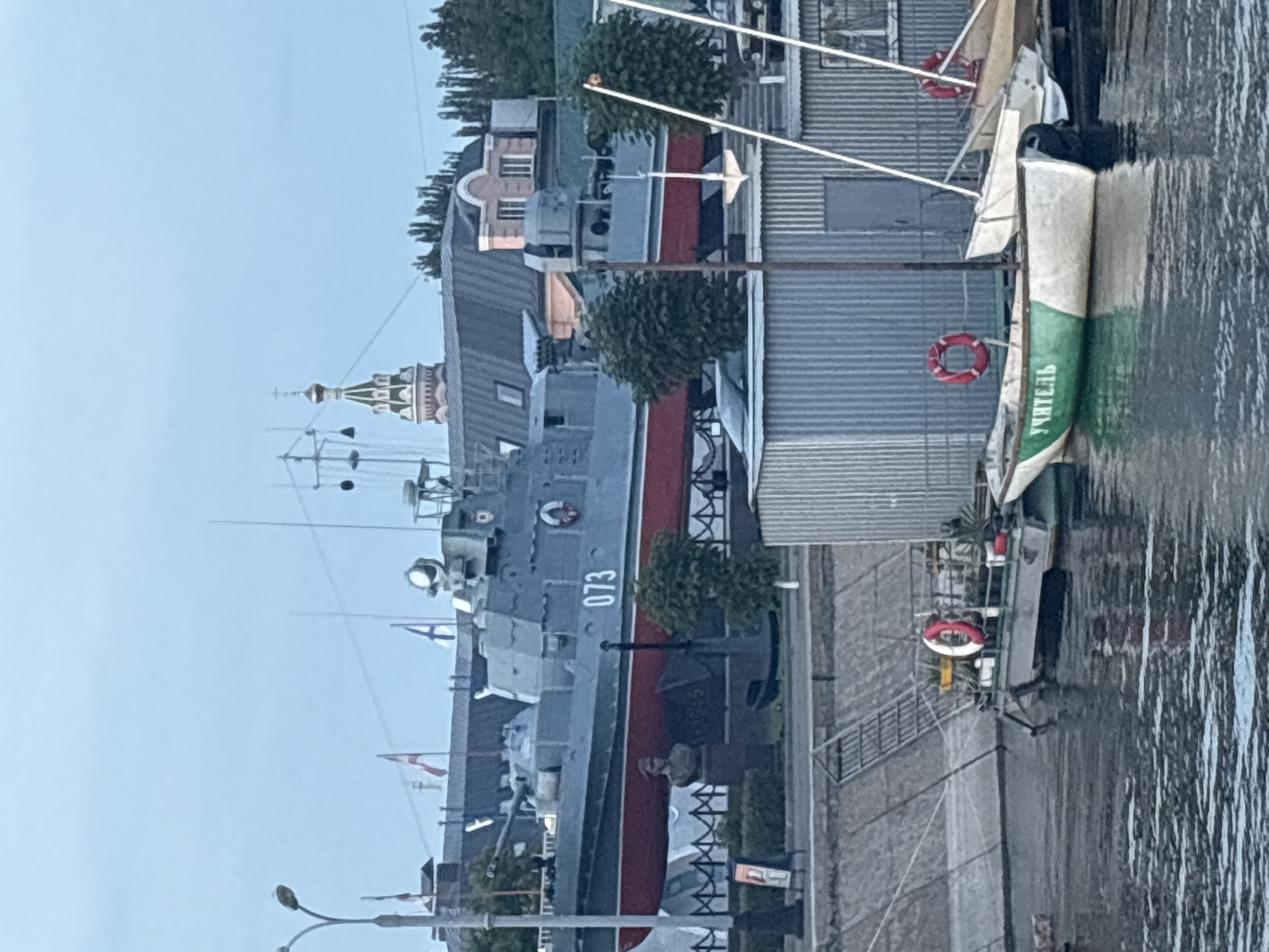 The image shows a view of a riverfront in Astrakhan, Russia, with a large grey and red ship docked and several smaller boats in the foreground. The ship, identified by the number "073" on its side, appears to be a former military vessel, possibly a torpedo boat, now serving as a museum or monument. A bust of a person stands near the ship. Behind the ship, buildings with distinctive architecture, including a domed church tower, are visible. In the foreground, two smaller boats are tied up. One is painted green and white with the word "Учитель" (Teacher) written on its side in Cyrillic. The scene takes place on a cloudy day, with the water of the river reflecting the sky and the boats. The riverbank is lined with a concrete embankment and some vegetation. A corrugated metal structure is located on the right side of the image, partially obscuring another boat.