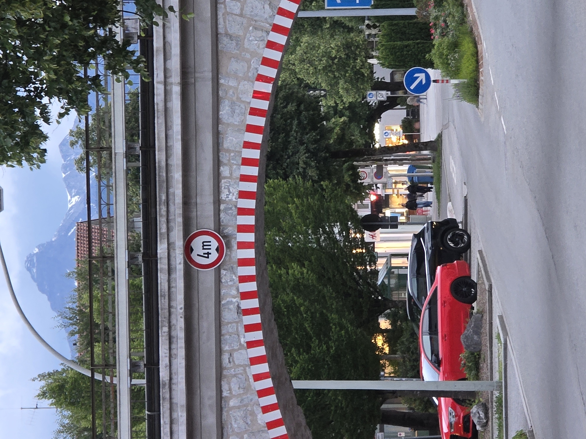 The image shows a street scene in Garmisch-Partenkirchen, Germany, under a stone archway with red and white striped markings. A sign on the archway indicates a height limit of 4 meters. Two cars are parked on the left side of the road, a red sedan and a black SUV. Several people are walking on the sidewalk in the background, and there are shops and trees lining the street. The sky is cloudy, with snow-capped mountains visible in the distance. A blue circular sign with a white arrow pointing to the right is visible on the right side of the road.