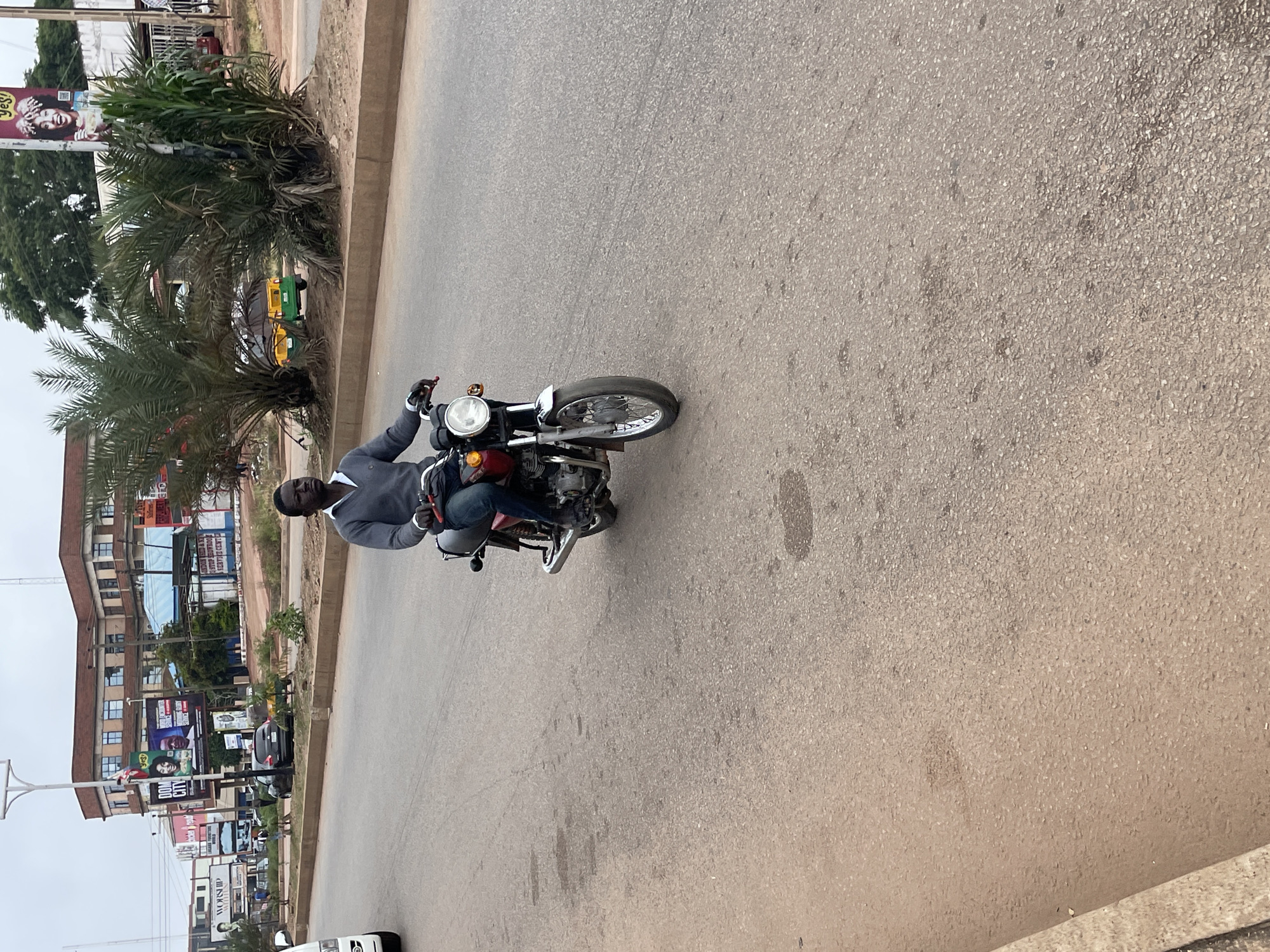 A man rides a red motorcycle down a paved street in Jos, Nigeria. He is wearing a grey sweater and blue jeans. The motorcycle has a silver bumper and a large headlight. In the background, there are buildings, palm trees, and a yellow and green auto-rickshaw. The sky is overcast.