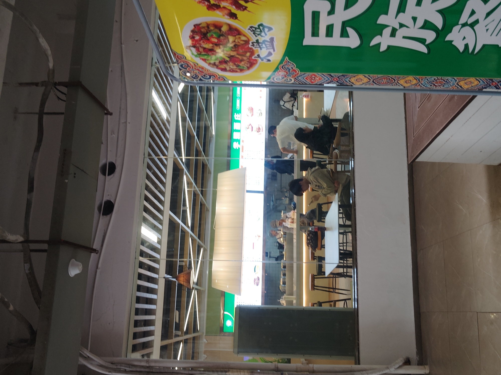The image displays an indoor food court or cafeteria area, likely located within a commercial building in Chongqing High-tech Zone, China. The perspective is from an upper level, looking down onto a dining area below, separated by a glass barrier.
**Subjects:** Several people are visible in the lower dining area, mostly seated at white tables with dark chairs or stools. They appear to be eating, waiting for food, or socializing. One person in a white shirt and dark pants is standing near what looks like a service counter. Another person in a light-colored jacket is seated, looking down. The reflections on the glass barrier also show distorted figures.
**Objects:** Prominently featured are large, colorful signs advertising food, particularly above the dining space. The area is brightly illuminated by long, rectangular fluorescent light fixtures. The infrastructure on the left includes exposed pipes and structural elements. The floor in the immediate foreground is tiled, while the lower dining level has a wooden or wood-patterned floor.
**Activity or event taking place:** People are engaged in typical food court activities such as dining, ordering meals, and conversing during what appears to be a mealtime.
**Notable details:** The scene suggests a modern, bustling commercial environment. The bright lighting indicates it could be daytime or an actively lit interior space. The architecture features an open layout with multiple levels.
**Visible text:** The most prominent text is on large banners. A yellow banner displays Chinese characters "新疆大盘鸡" (Xīnjiāng Dàpánjī - Xinjiang Big Plate Chicken) and "烤肉" (kǎoròu - roasted meat/kebab), accompanied by images of the dishes. Below this, a large green sign prominently features white Chinese characters, reading "民族美食" (Mínzú Měishí - Ethnic Delicacies), suggesting a restaurant specializing in diverse regional cuisines. A smaller green illuminated sign also shows "米线" (Mǐxiàn - Rice Noodles) and "套餐" (Tàocān - Set Meal).