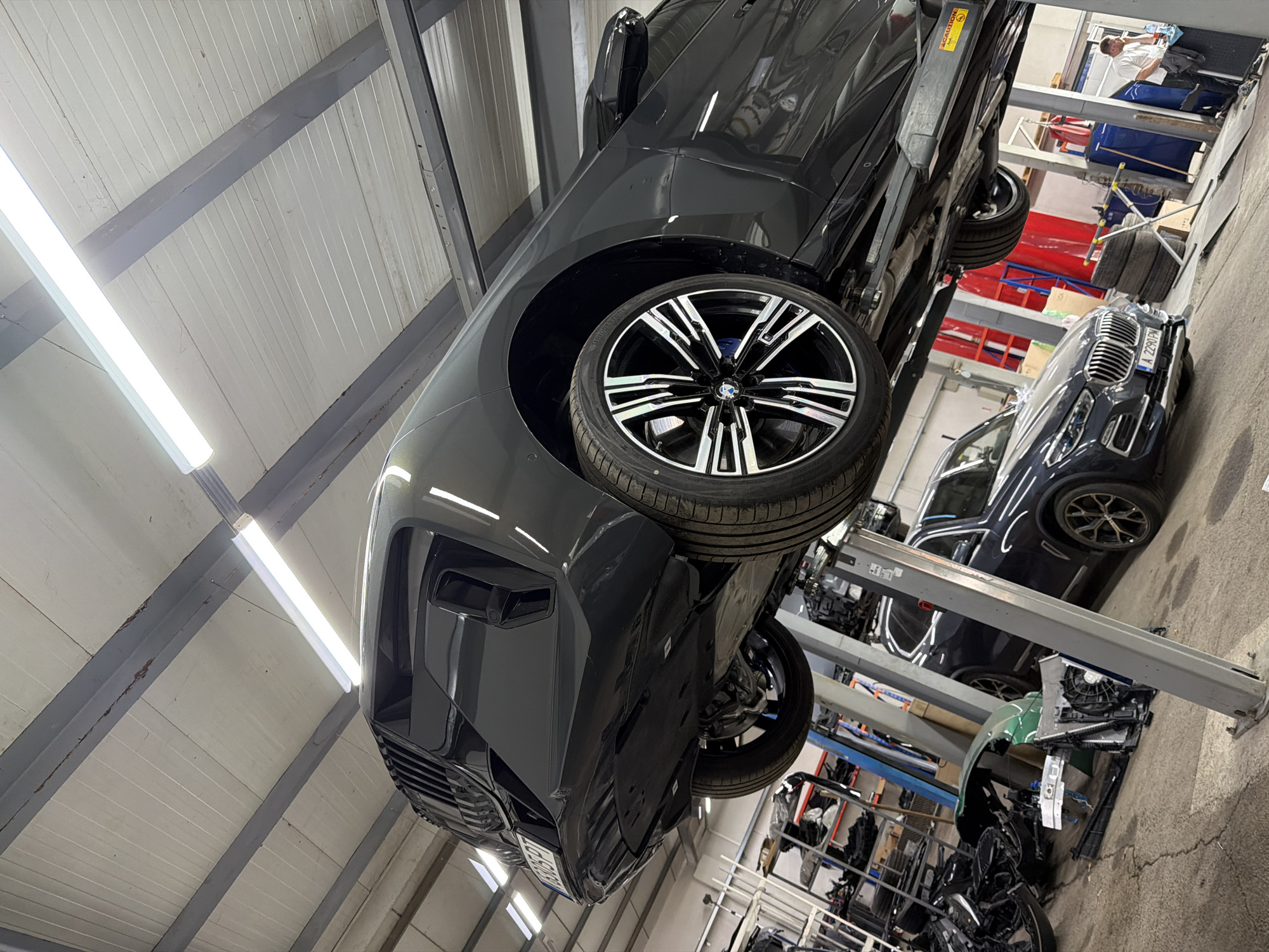 The image depicts the interior of an auto repair shop or garage, with a dark grey or black SUV prominently elevated on a hydraulic car lift, dominating the left and center of the frame. The vehicle, possibly a BMW given the distinctive wheels with a visible BMW logo and blue brake calipers, is positioned with its front end facing towards the bottom left. Its undercarriage is visible, and the front bumper is angled towards the viewer.
In the background, to the right, another dark-colored car, also appearing to be a BMW, is parked on the concrete floor. This car has a European license plate that reads "A 2290 PM". Further in the background, a person wearing a white shirt can be seen standing and appearing to work at a workbench or service area.
The garage features an industrial aesthetic with a high ceiling composed of white corrugated metal panels and exposed grey support beams. Bright fluorescent lights illuminate the space. Various car parts and equipment, including spare tires, shelving units, and what appears to be a disassembled front end of a vehicle (possibly green in color) with a radiator, are scattered around the concrete floor, indicating ongoing maintenance or repair work. A "CAUTION" sticker is visible on the structure of the car lift. The overall scene suggests a busy and functional automotive service environment in Burgas, Bulgaria, given the license plate format.