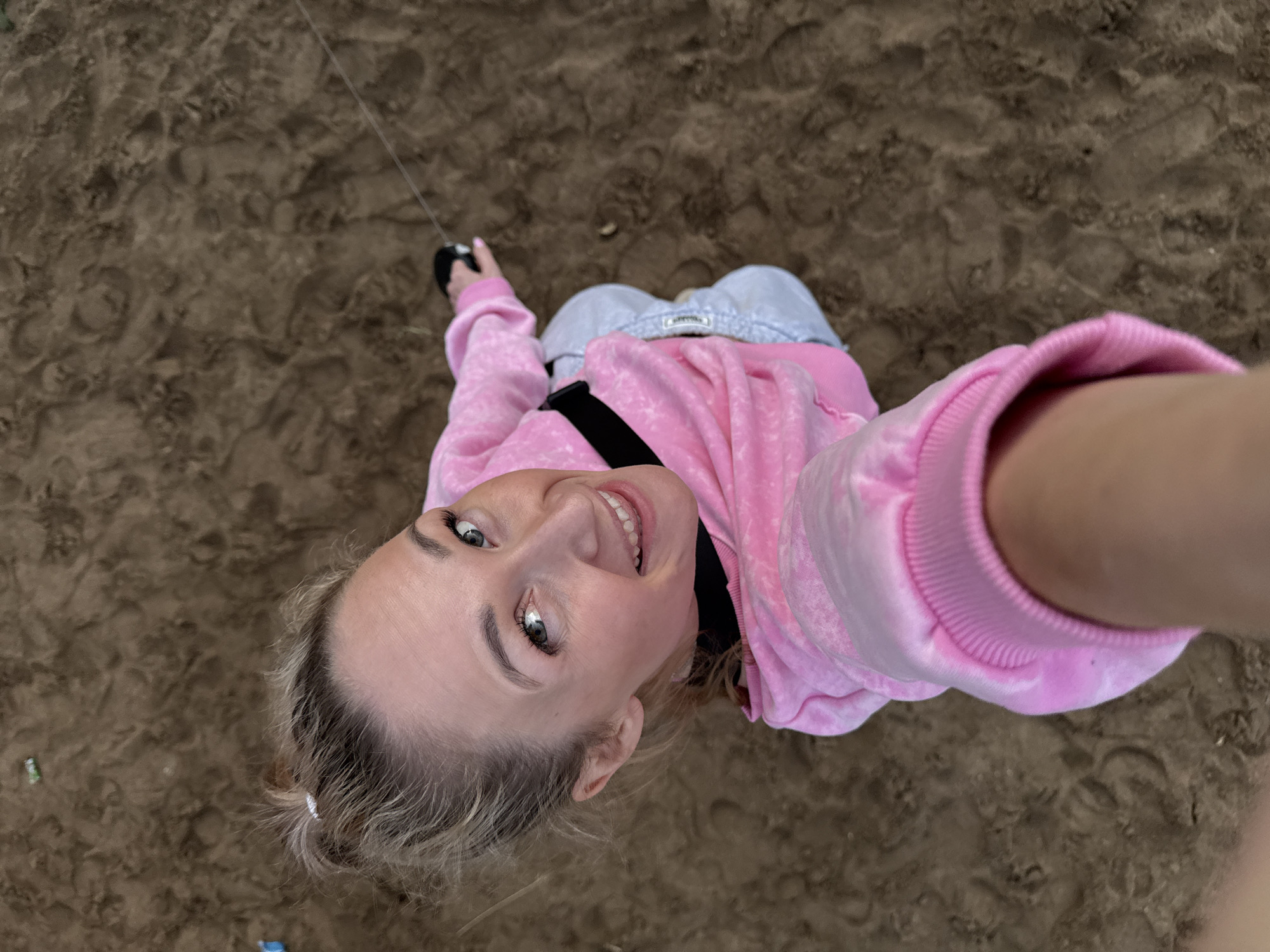 In a top-down shot, a woman with blonde hair tied in a messy bun smiles at the camera. She is wearing a pink tie-dye sweatshirt and light blue sweatpants. Her left hand is visible, holding what appears to be kite string. The background consists of a sandy surface with footprints. The scene is likely outdoors, possibly in a park or on a beach. The image was taken in the city of Chaikovsky, Russia.