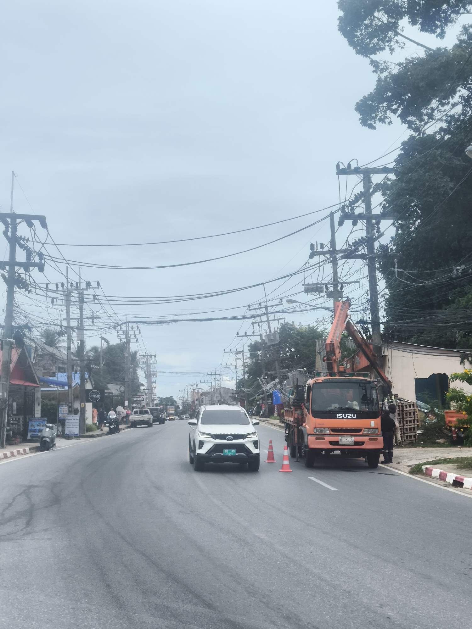 The image depicts a street scene in Baan Chaweng Noi, Thailand, on an overcast day. A white SUV is in the foreground on the right, traveling on a wet asphalt road. To the left of the SUV, an orange Isuzu truck with a crane is parked, with two individuals standing beside it. Three orange traffic cones are placed in front of the truck. A line of utility poles with numerous wires runs along both sides of the road, characteristic of the area. Buildings with awnings and signs line the street on the left. Further down the road, other vehicles and motorcycles are visible, as well as more utility poles and foliage. The overall impression is a typical street scene with ongoing infrastructure work or maintenance.
